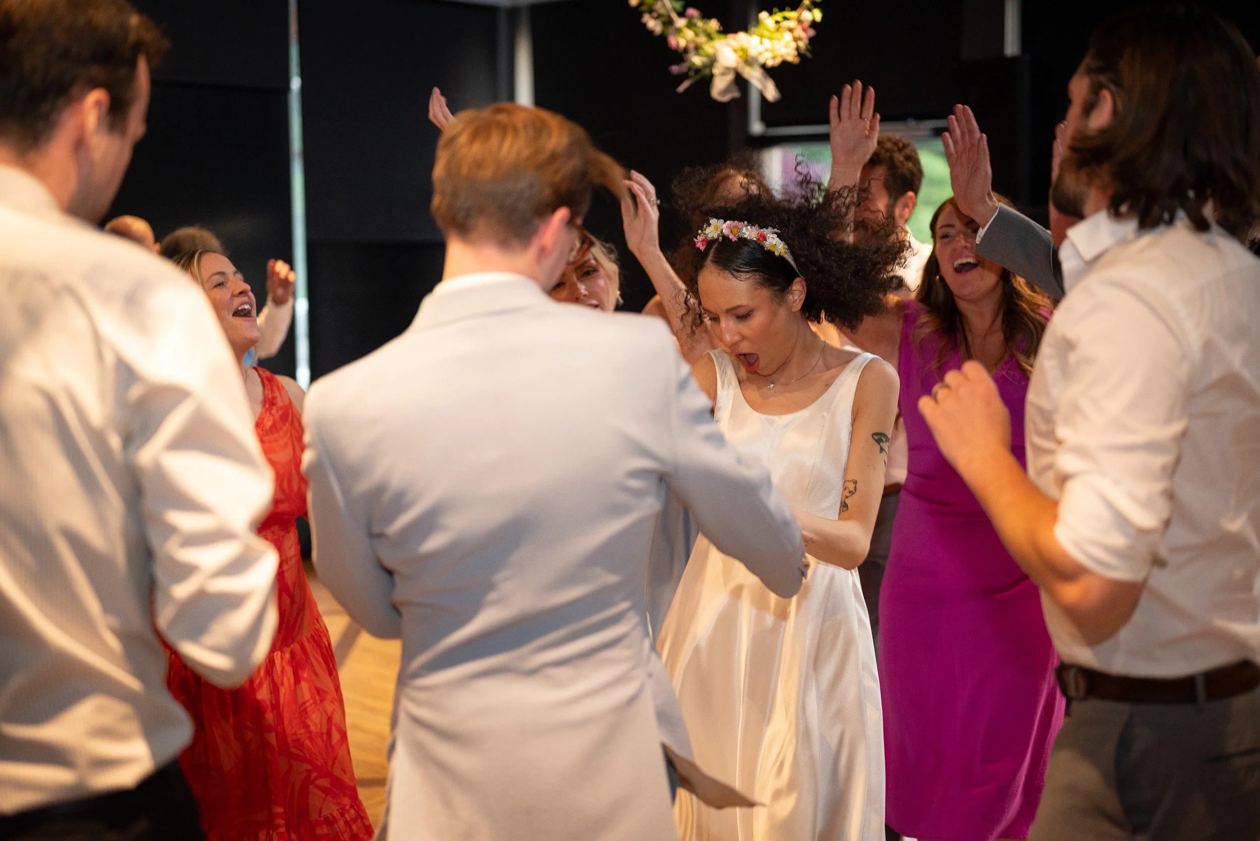 Group of people dancing in a circle at a celebration, with some raising hands and smiling; woman in the center wearing a flower crown and white dress.