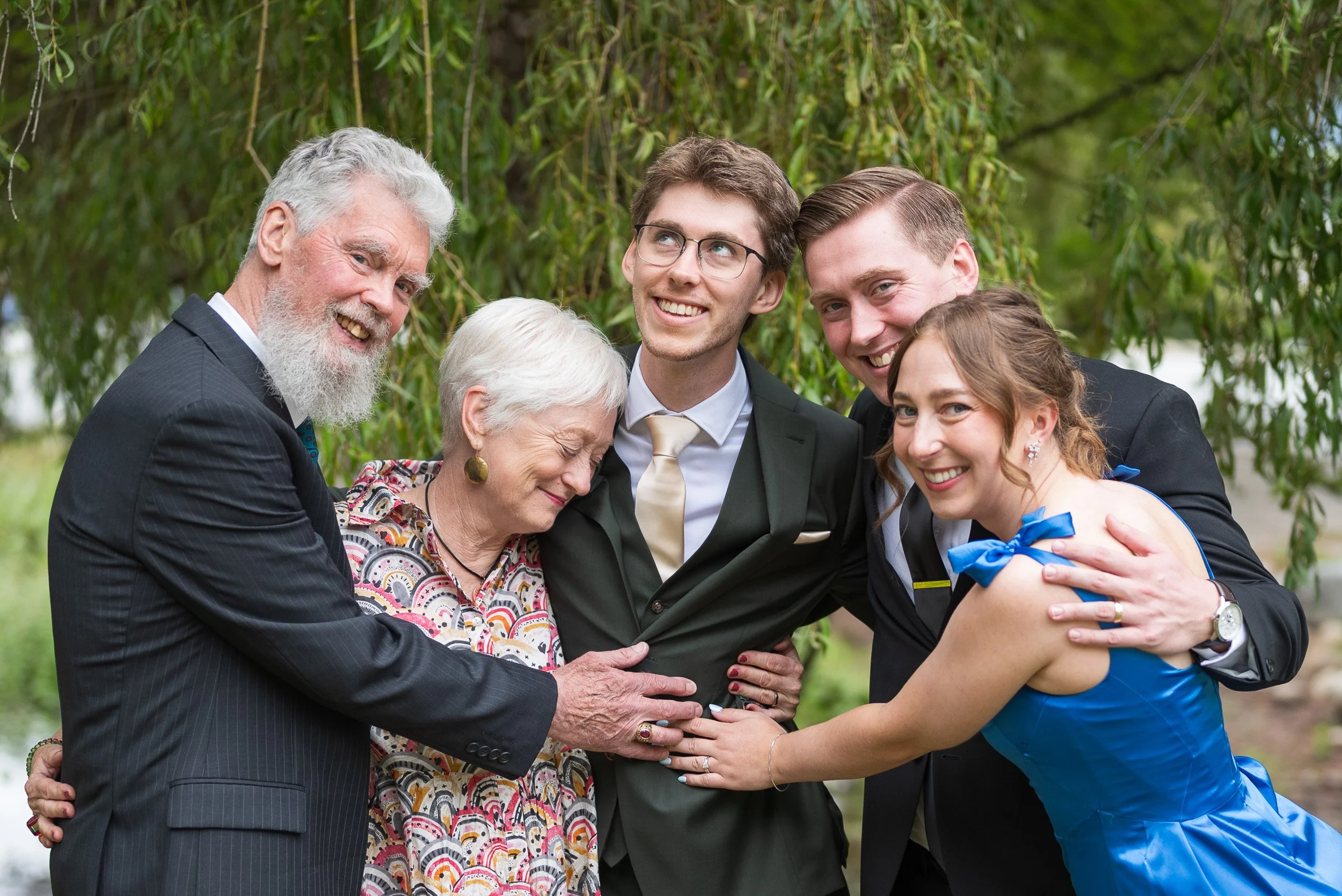 A group of six people, including an elderly couple, two young men, and a young woman in a blue dress, smiling and hugging in a park with green foliage in the background.