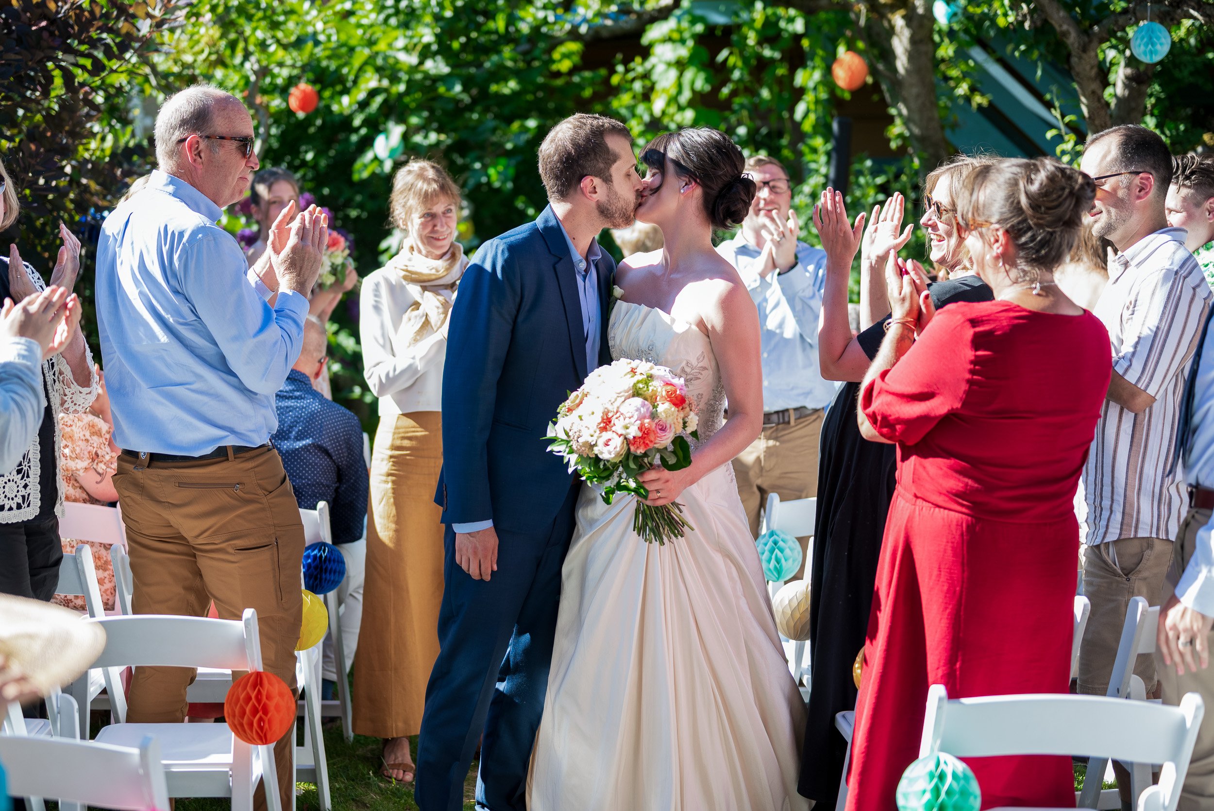 A newlywed couple shares a kiss during their outdoor wedding ceremony, surrounded by family and friends, with colorful decorations and lush greenery in the background.