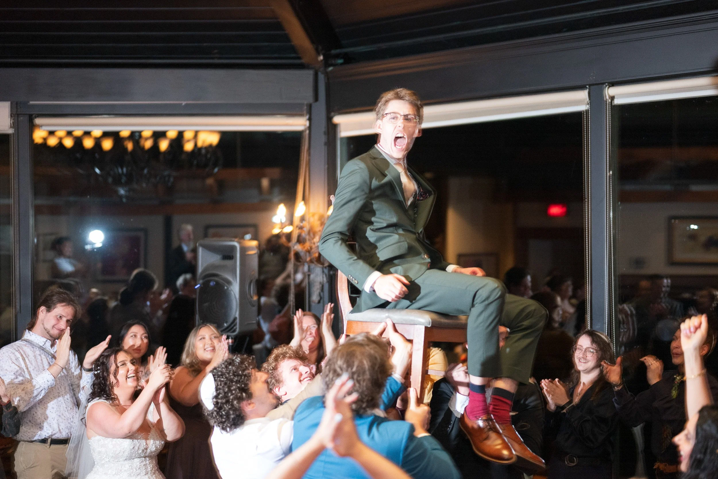 A man in a suit and glasses is sitting on a chair, on a platform, with his mouth open and a surprised or excited expression. He is surrounded by a crowd of smiling and clapping people in an indoor venue.