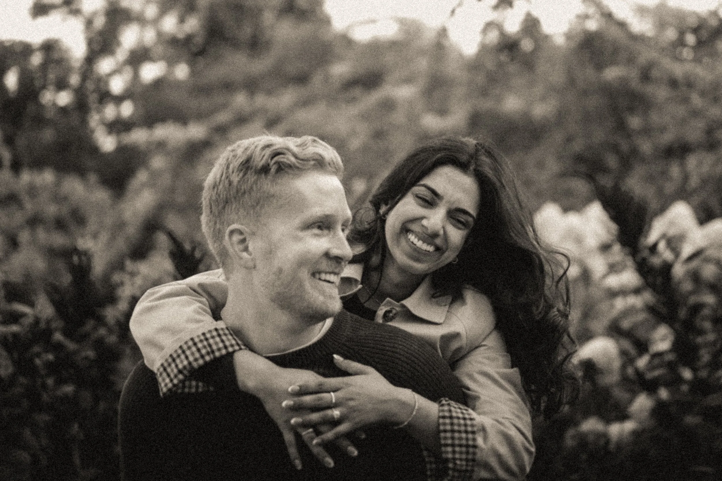 An artistic engagement black and white photo of a man and woman smiling warmly, embracing outdoors with trees in the background.
