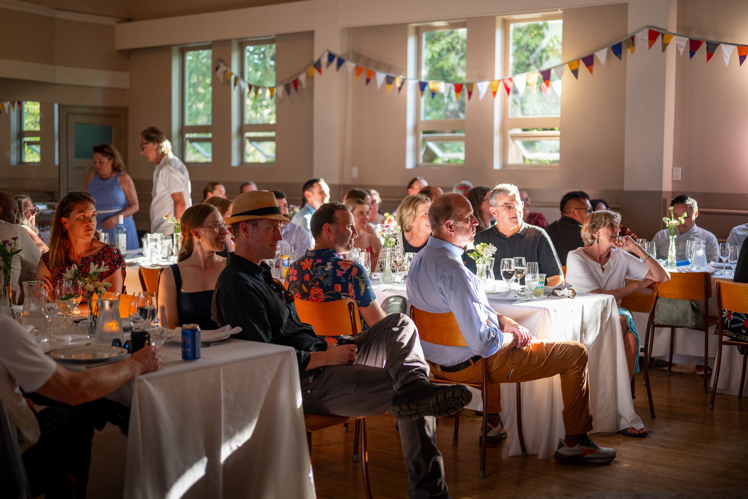 Guests seated at tables during a social gathering or celebration in a well-lit room with large windows and colorful bunting.