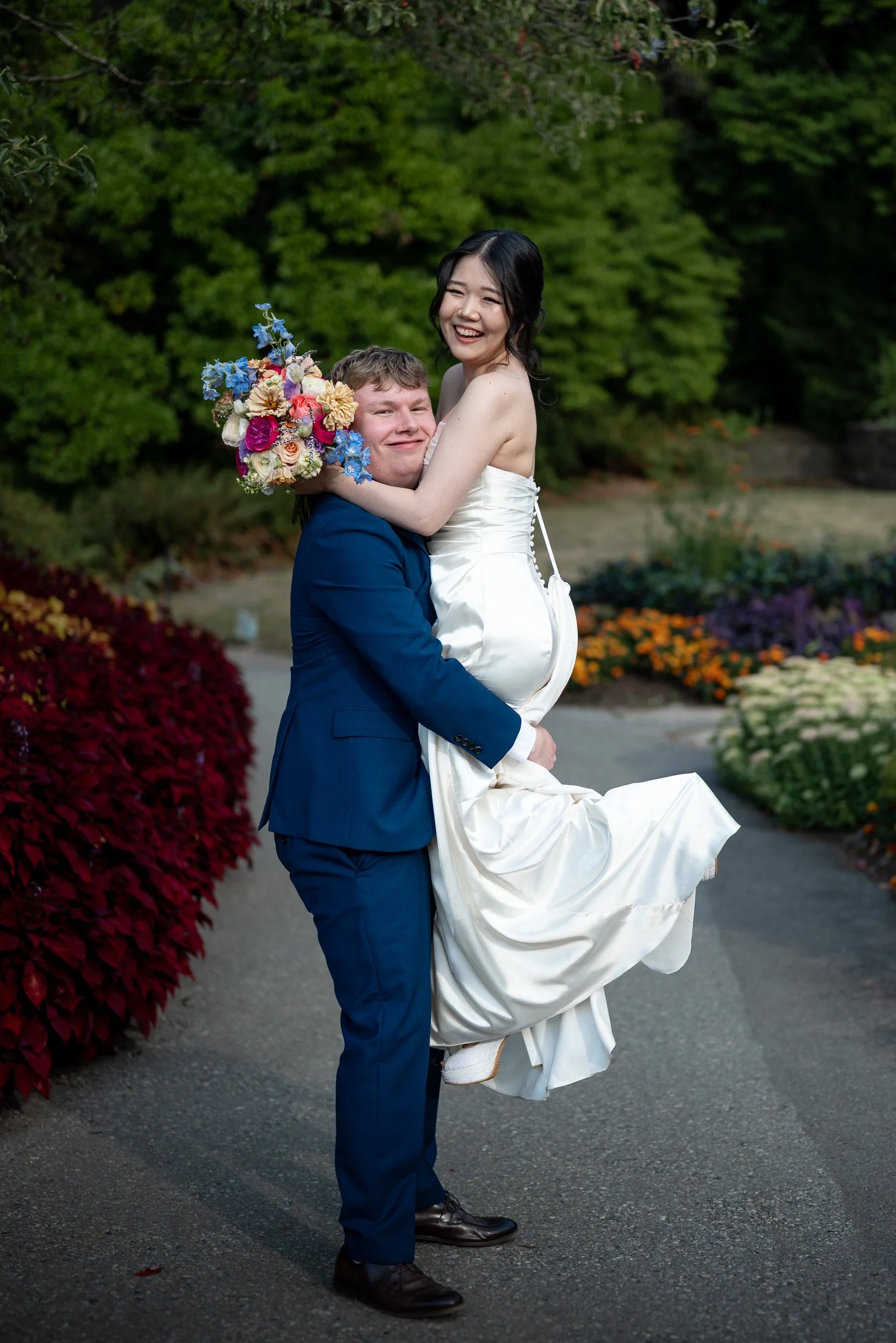 A man in a blue suit holding a woman in a white wedding dress and holding a colorful bouquet of flowers, standing outdoors with green trees and colorful bushes in the background.