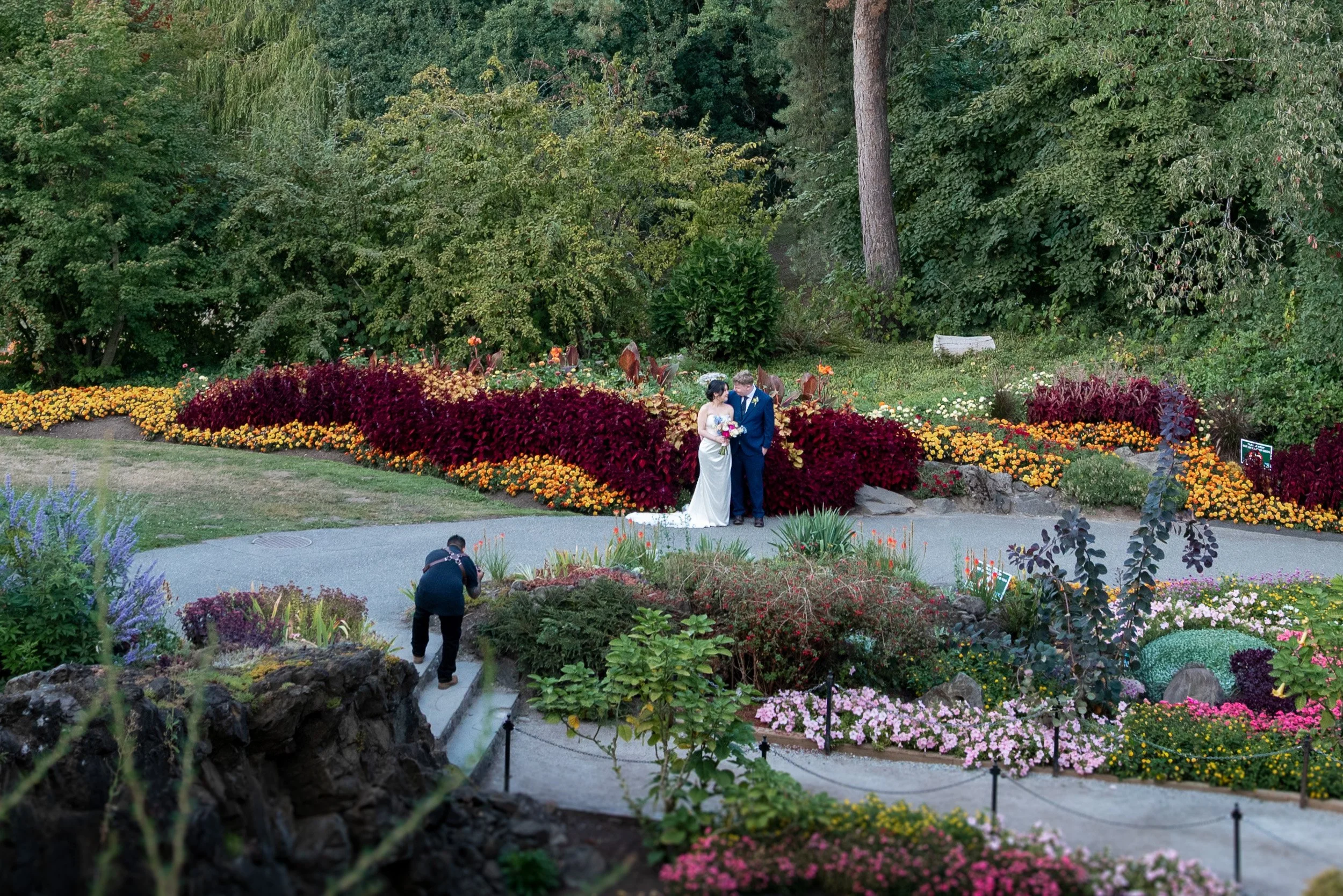 A wedding couple standing together in a lush garden filled with colorful flowers and green trees, with a photographer taking pictures and a person walking nearby.