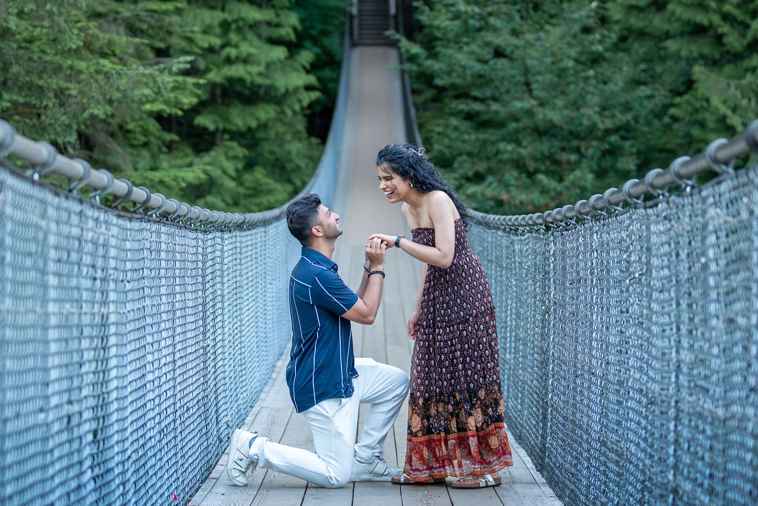 A man kneeling on one knee proposing to a woman on Capilano Suspension bridge surrounded by greenery, both smiling.