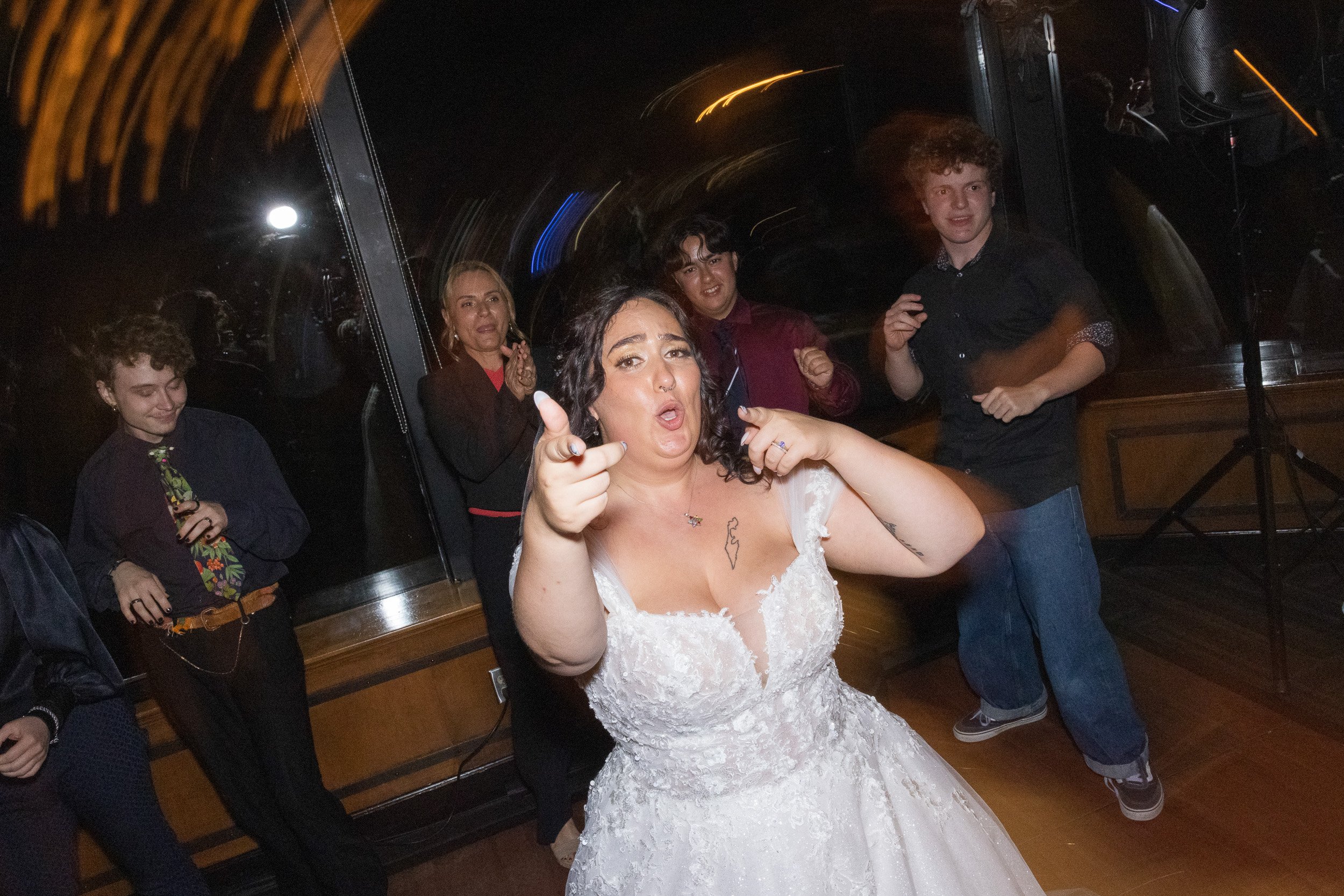 A woman in a wedding dress making a playful gesture with her hands in front of a group of people at a celebration or party.