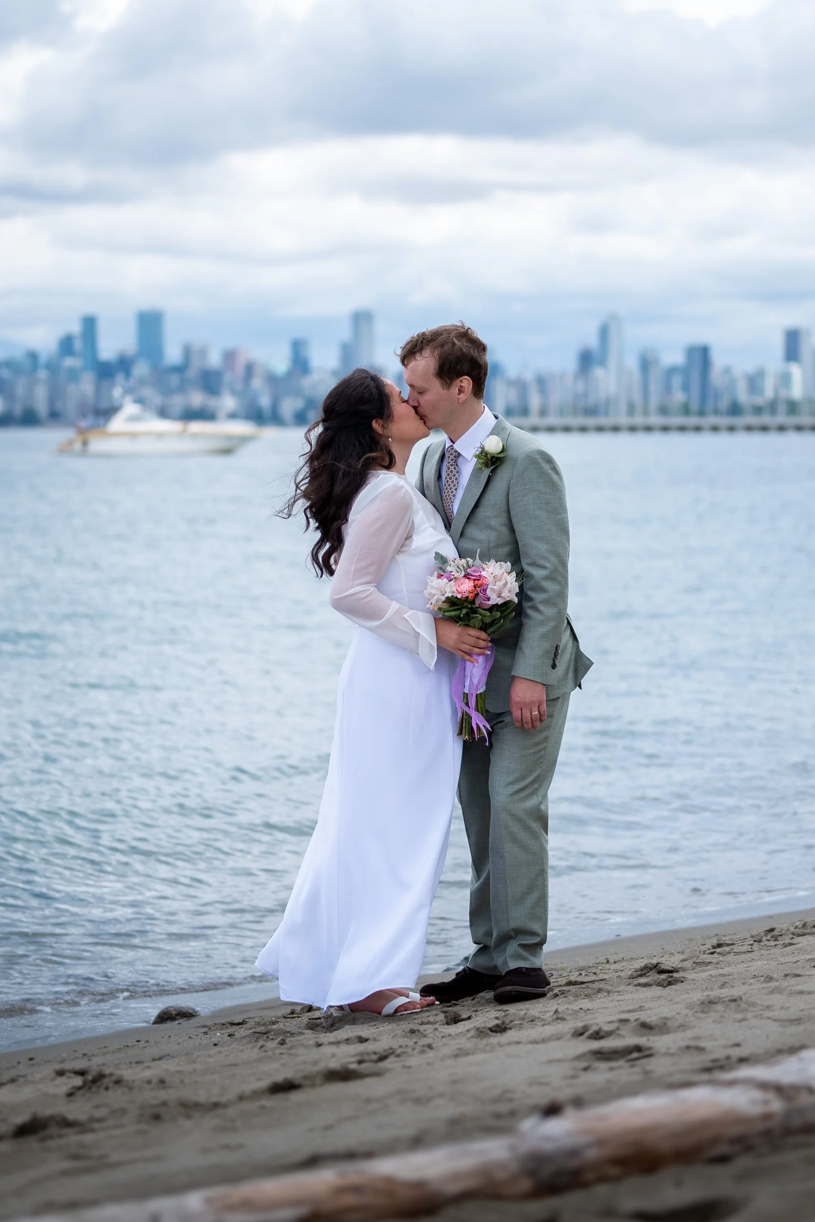 A newlywed couple sharing a kiss on Jericho Beach in Vancouver, with city skyline and a boat in the water in the background.
