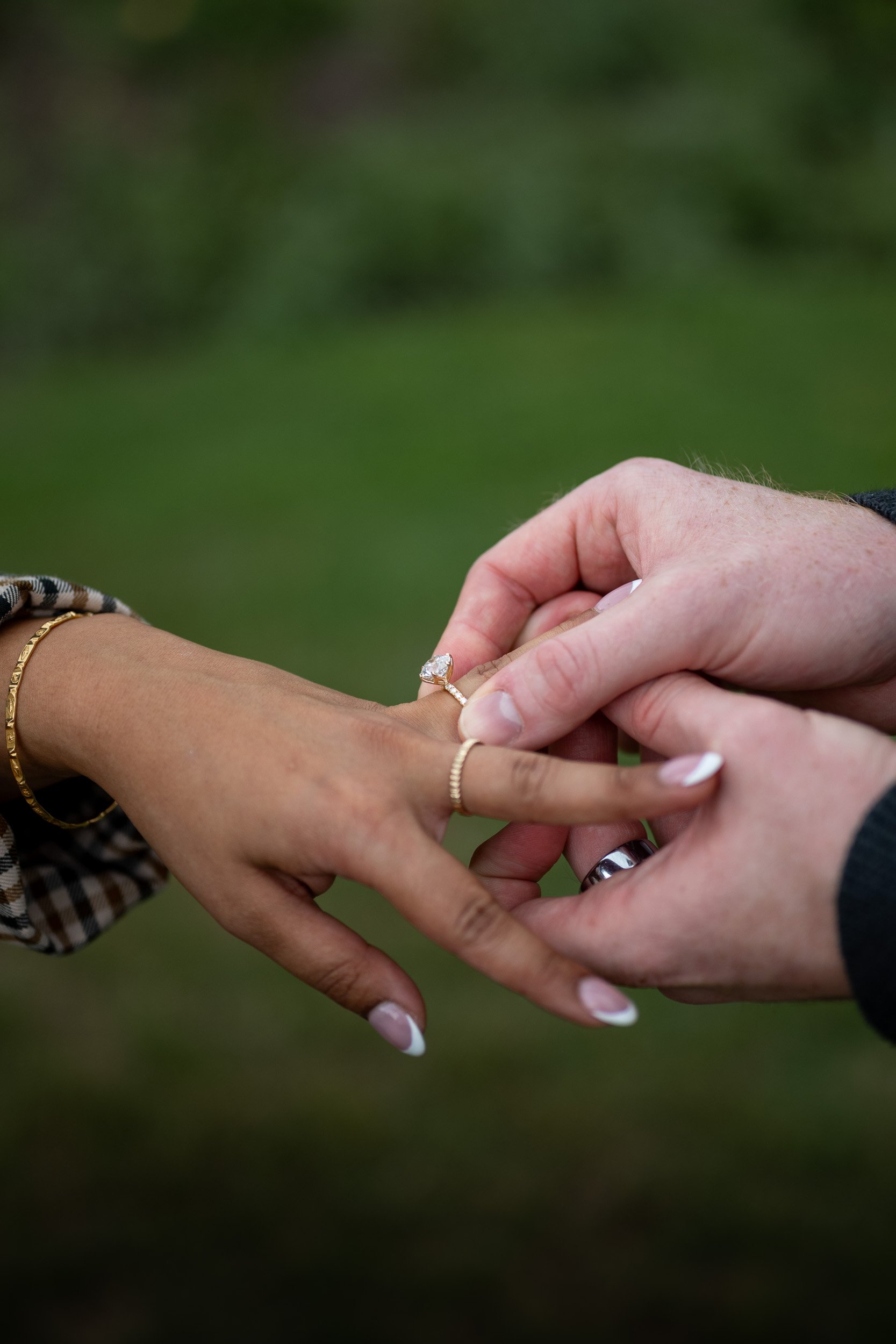 Close-up of three hands, one putting a ring on the ring finger of another, with a green blurred background.