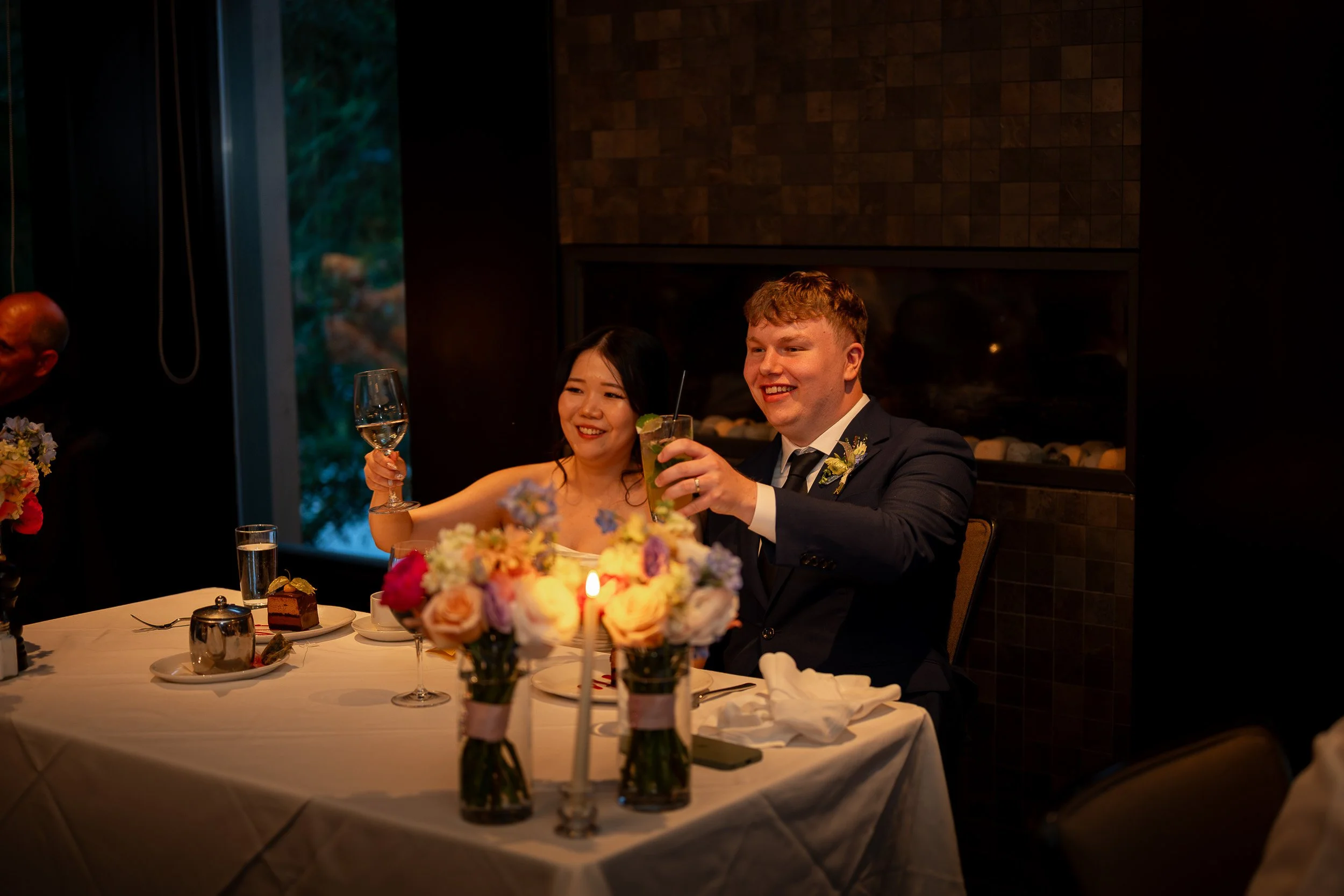 A newlywed couple at a wedding reception sitting at a decorated table with flowers, desserts, and drinks, smiling and raising their glasses.