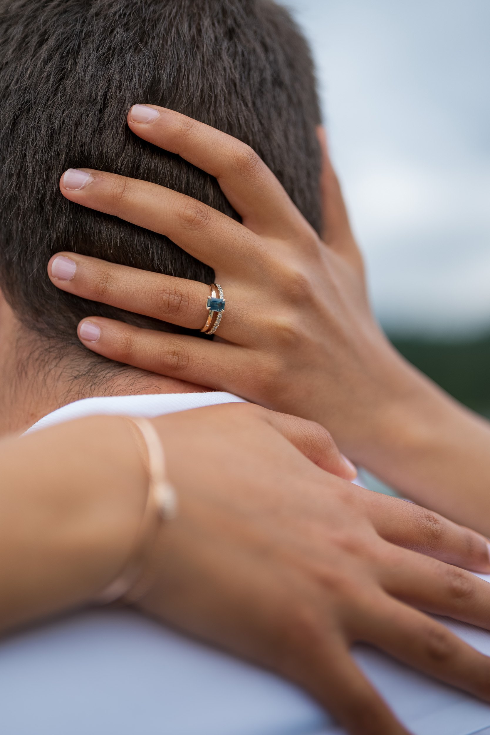 Close-up of a person's hand with a ring on the ring finger, gently touching the back of their head and neck. The background is blurred.