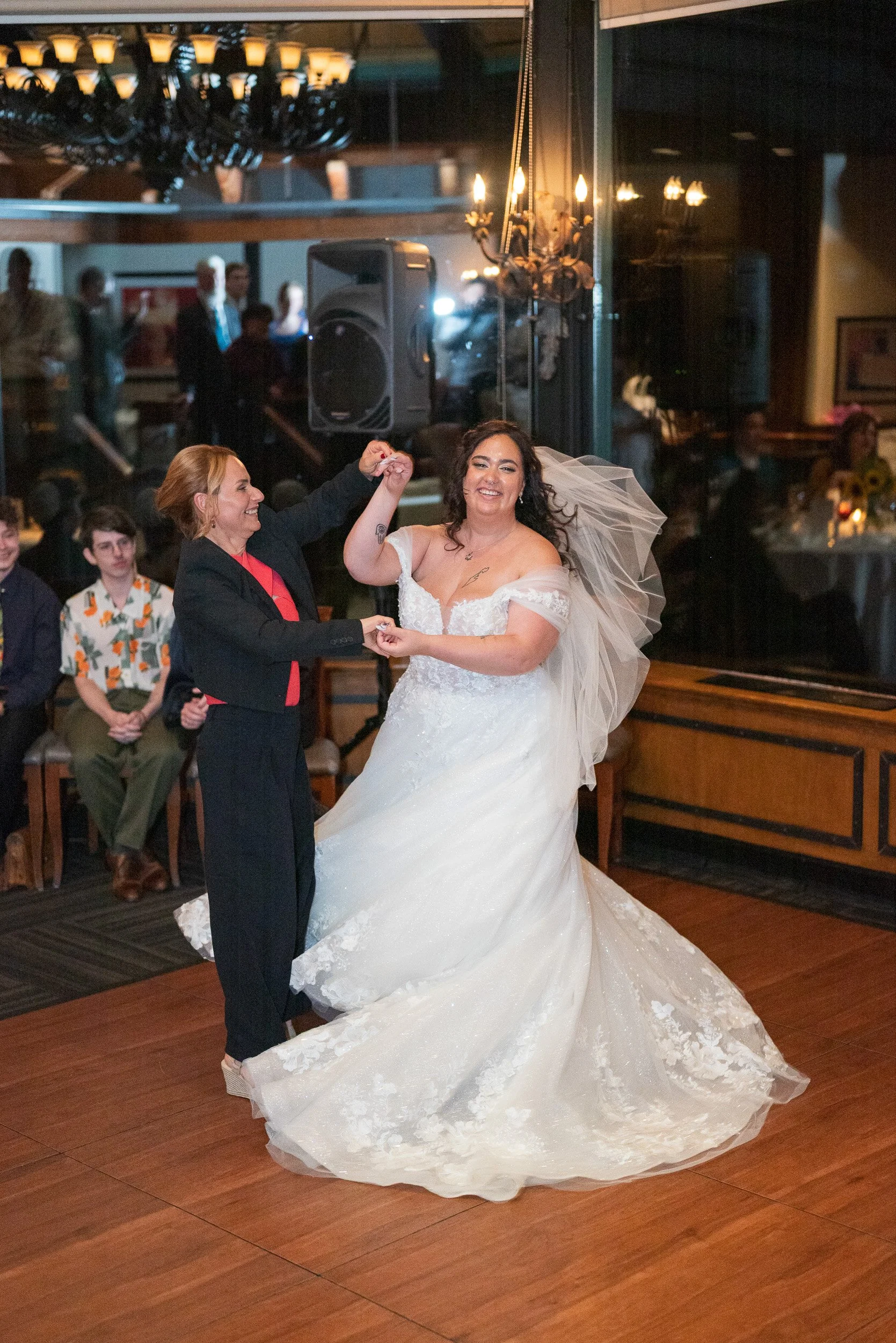 Two women, one in a wedding dress and the other in a suit, dancing at a wedding reception with guests watching in the background.