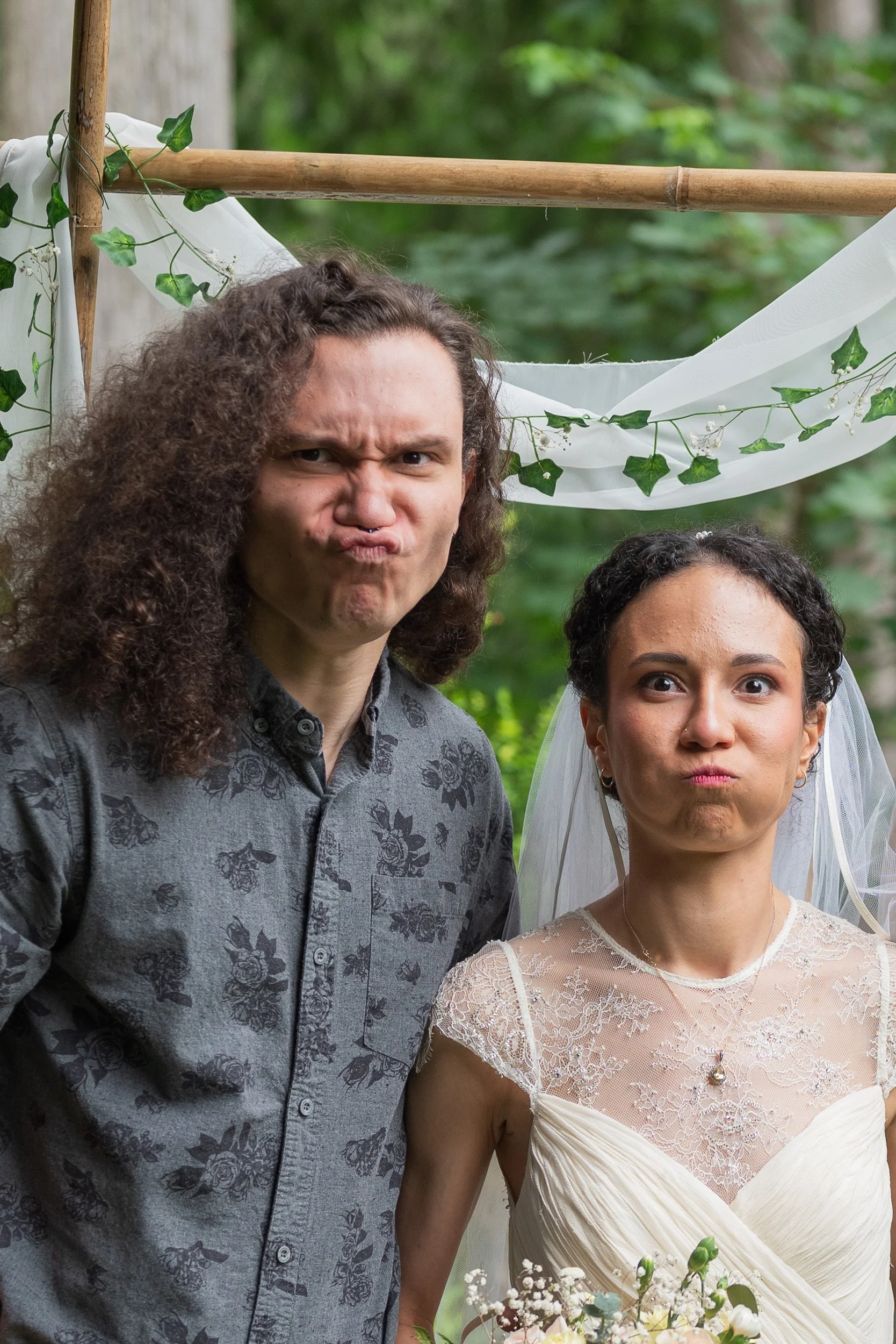 A man with long curly hair making a funny face and a woman in a wedding dress with a veil, standing outdoors surrounded by greenery and wedding decorations.
