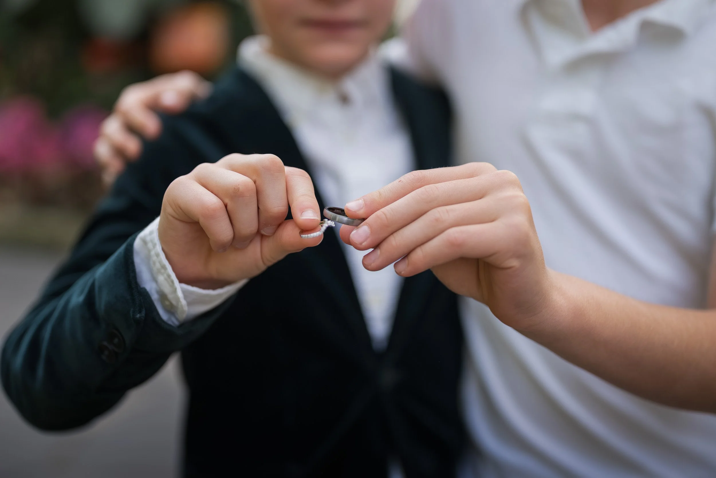 Person in a dark jacket and white shirt holding an engagement ring while another person in a white shirt stands nearby, with a blurred background.