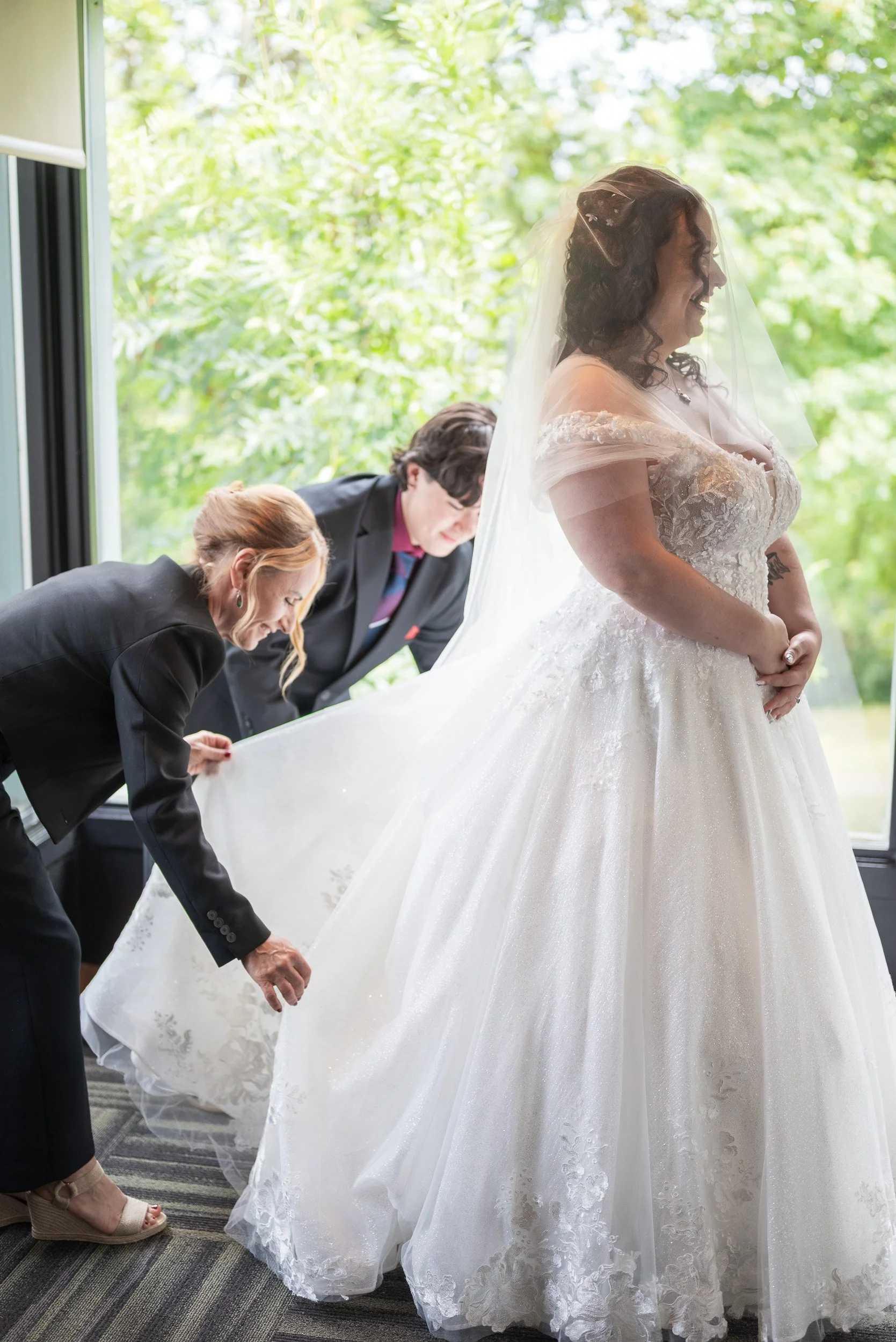 A bride in a lace wedding gown and veil standing by a window, smiling, with two women adjusting her dress, these are getting ready photos at Seasons in the Park restaurant in Queen Elizabeth Park.