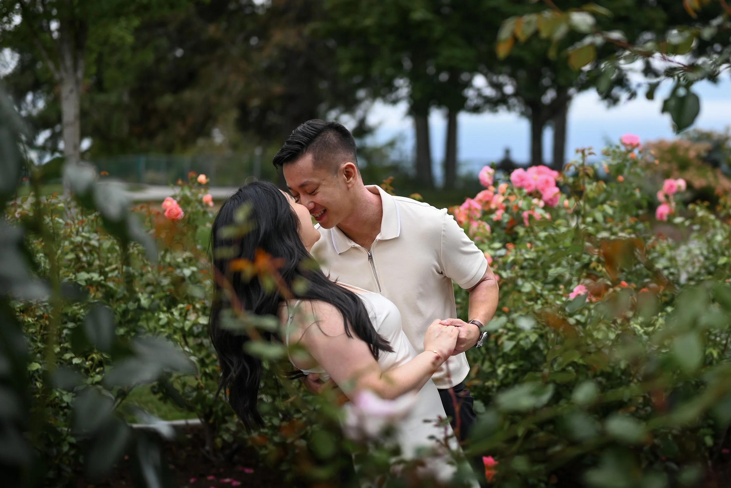 A couple sharing a kiss and holding hands in a garden full of pink roses with trees in the background at Burnaby Mountain.