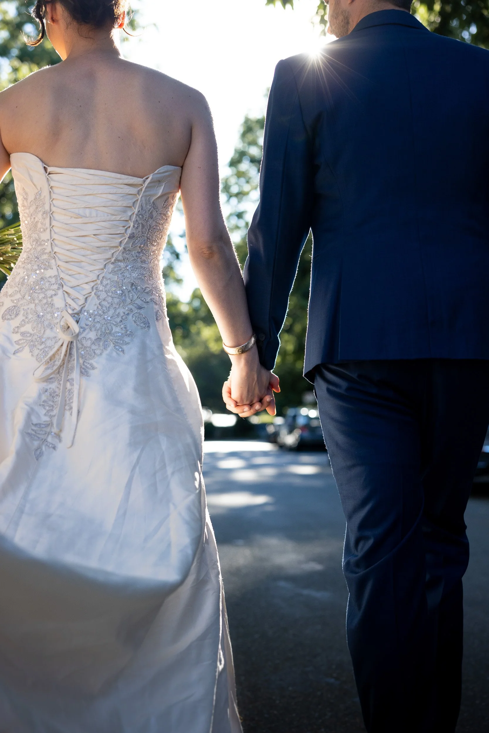 A bride and groom holding hands outdoors during sunset, with sunlight shining behind them, wearing wedding attire.