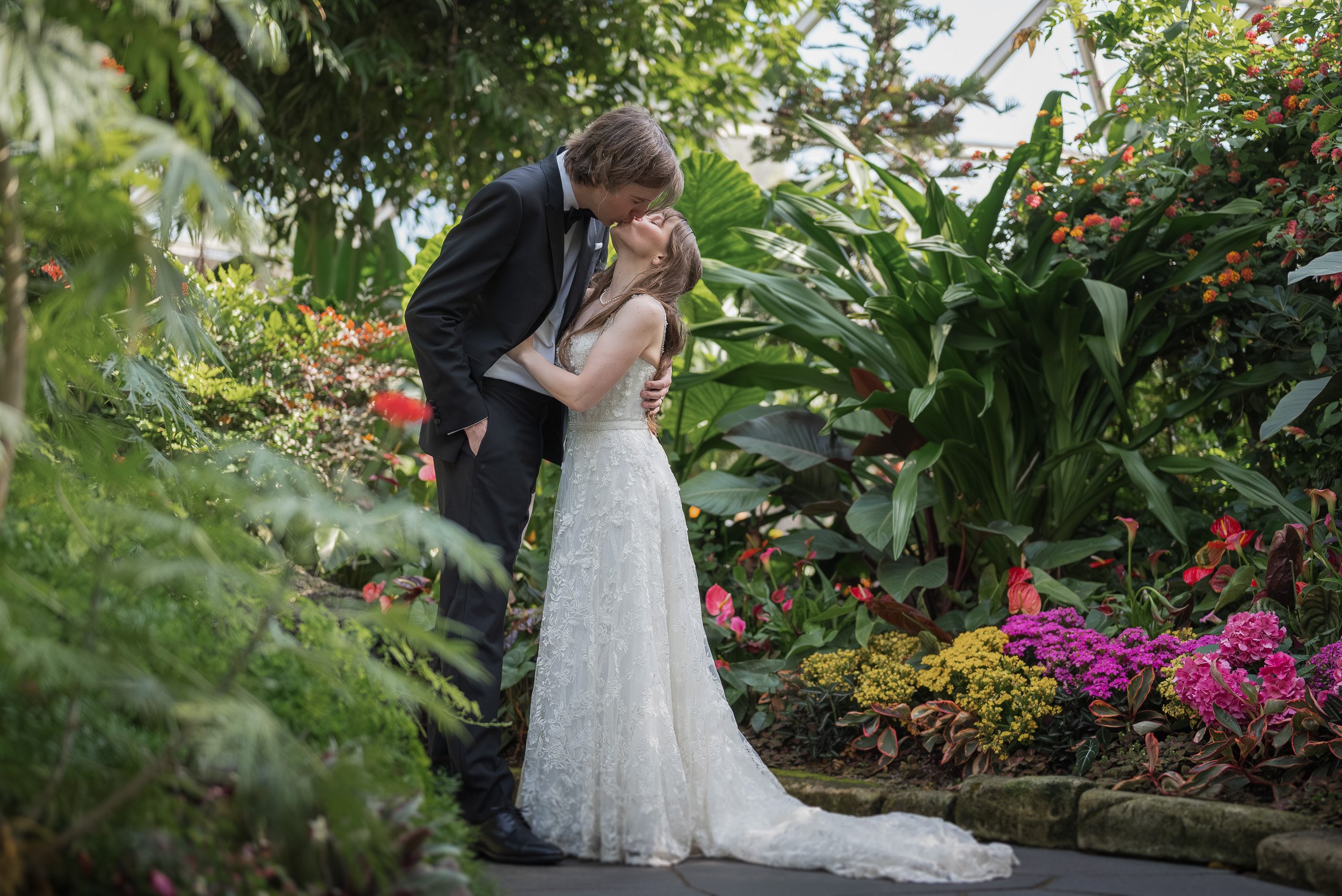 A bride and groom kissing in a lush garden filled with colorful flowers and green foliage.