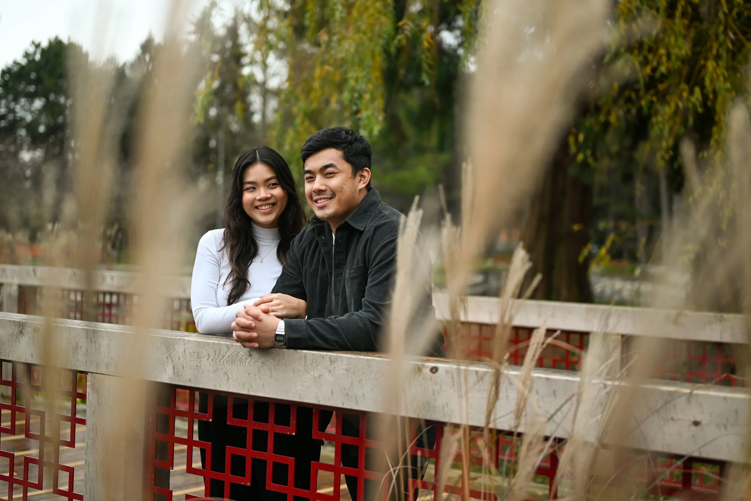 A smiling young woman and man sitting at a wooden fence outdoors, surrounded by trees and grass, with tall dry grasses in the foreground.