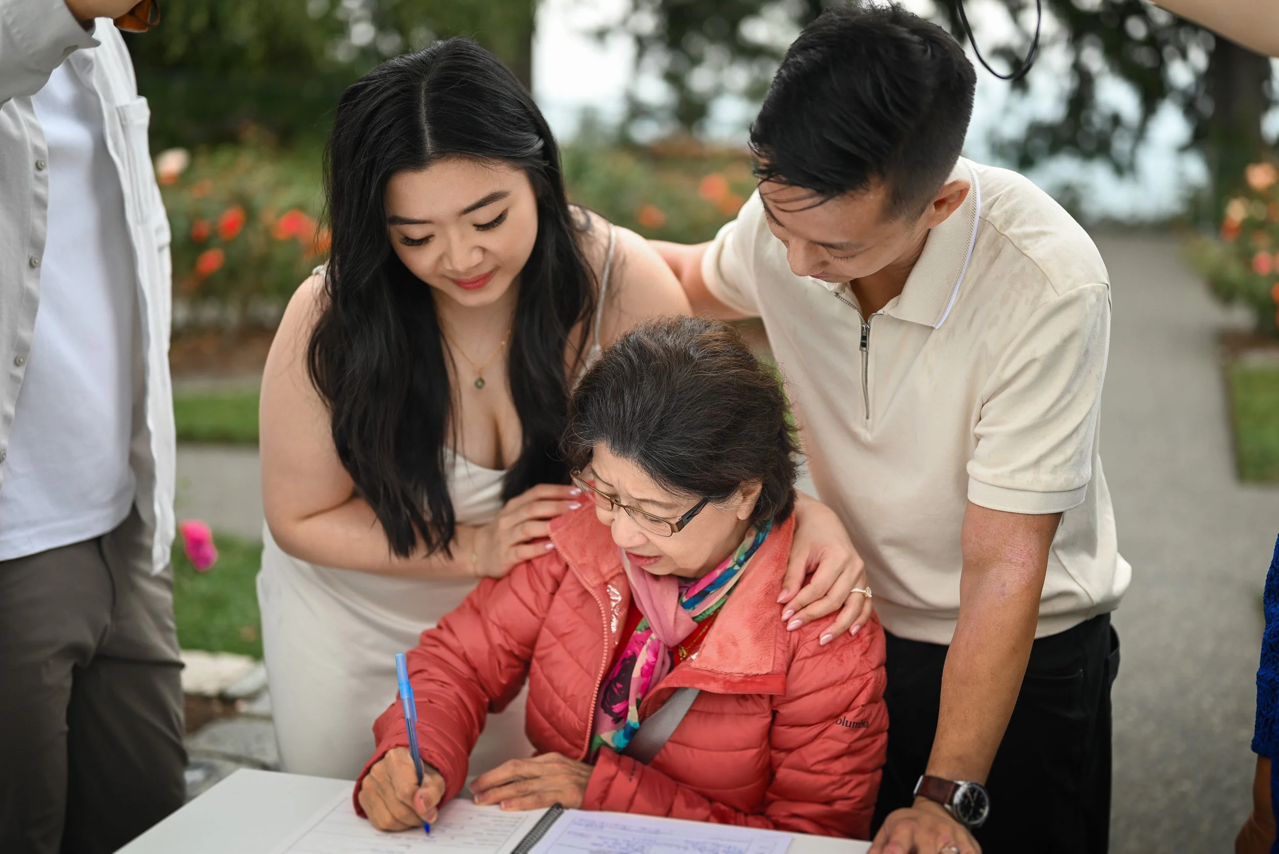 A group of people, including an elderly woman, is gathered around a table outdoors. The elderly woman is sitting and writing in a notebook with a pen, and others are standing around her, looking at the notebook. The scene appears to be friendly and s