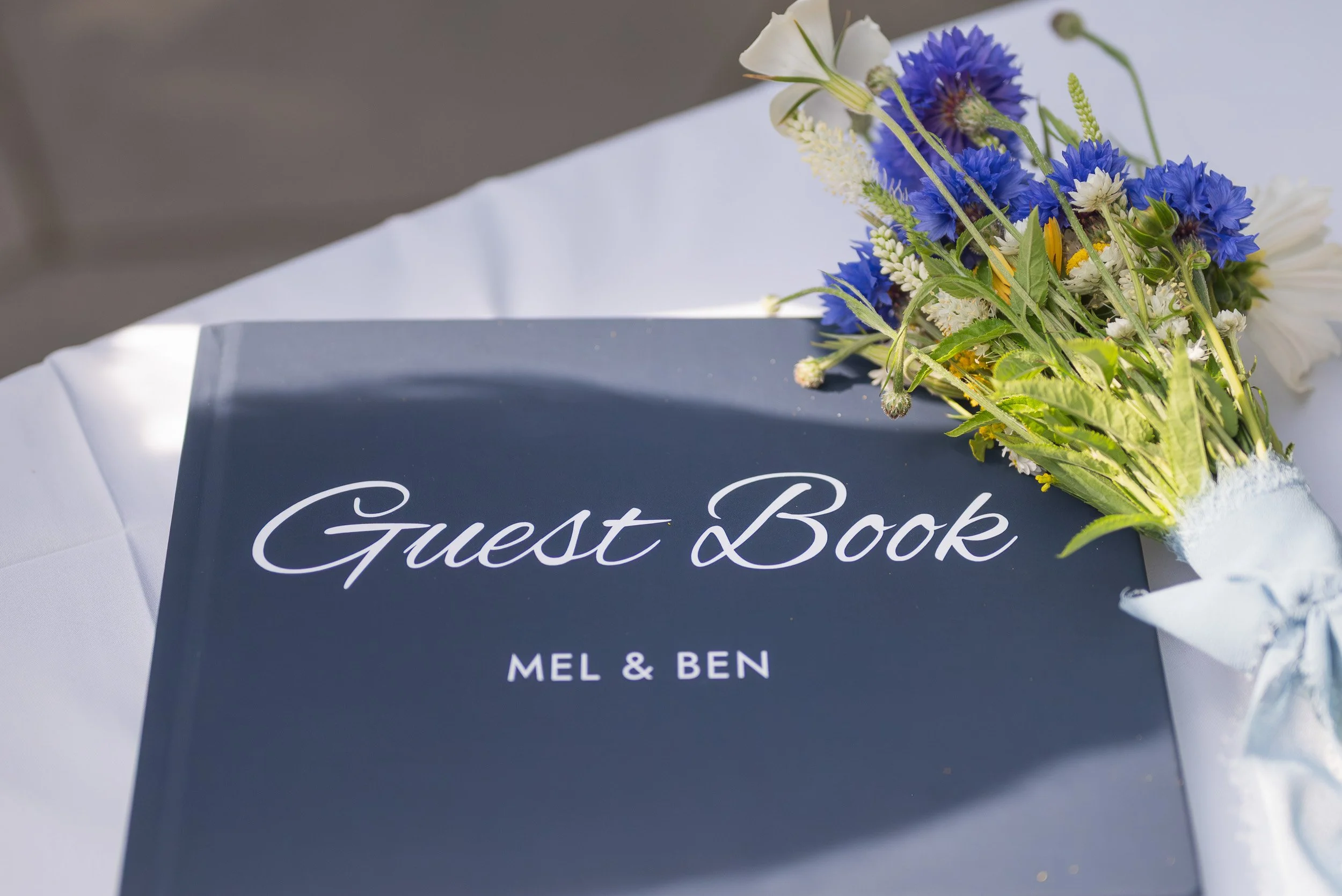 A guest book with a floral arrangement on top, placed on a white tablecloth. The guest book is dark-colored with white cursive writing that says 'Guest Book' and names 'Mel & Ben' underneath.