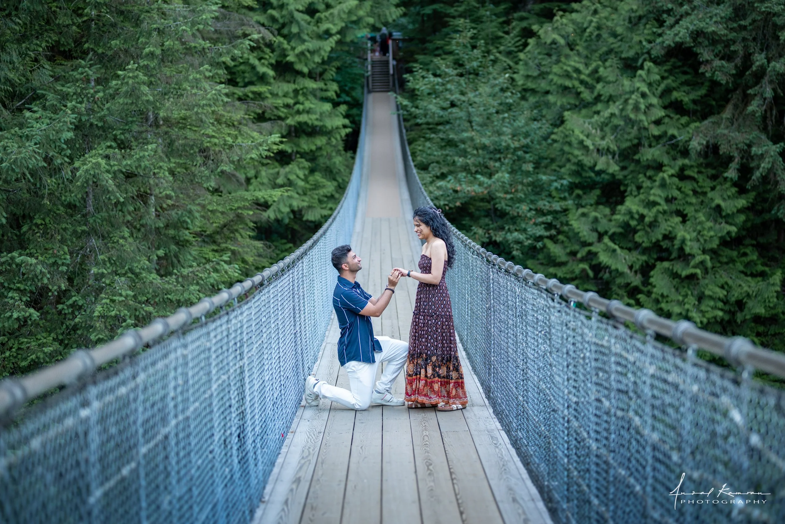 A man proposing to a woman on Capilano suspension bridge surrounded by dense green forest. The man is kneeling on one knee, holding the woman's hand, who is standing and smiling.
