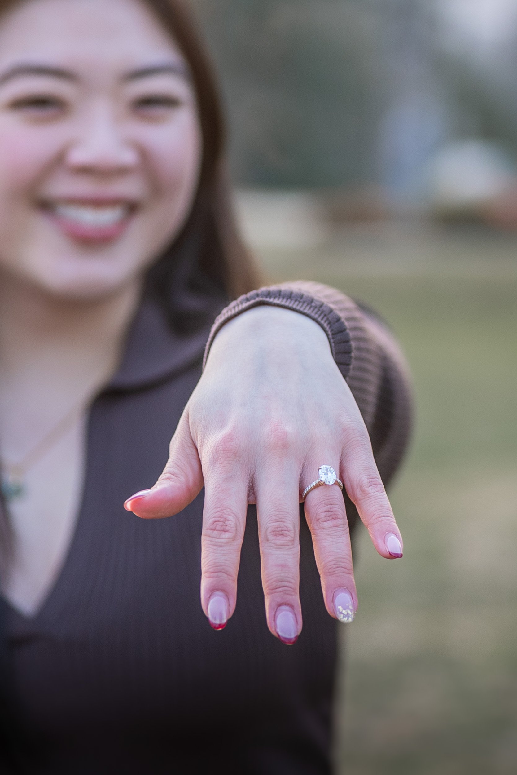 Close-up of a woman showing off her left hand with a large diamond engagement ring on her ring finger, with her smiling face slightly blurred in the background.