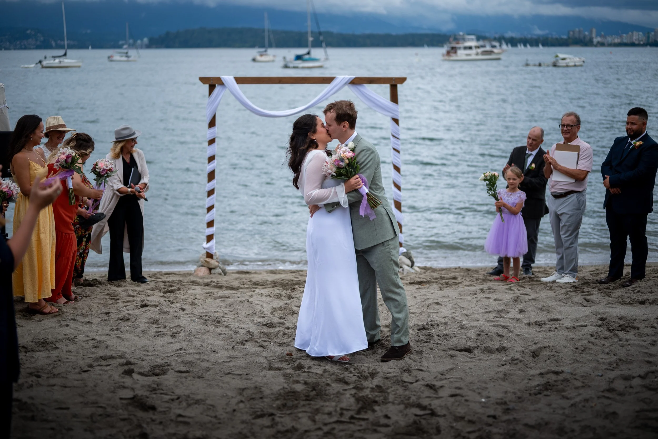 A couple kisses during their outdoor wedding ceremony on Jericho beach with boats on the water in the background. Guests stand on each side of the couple, holding flowers and clapping, with a wooden arch decorated with white fabric.