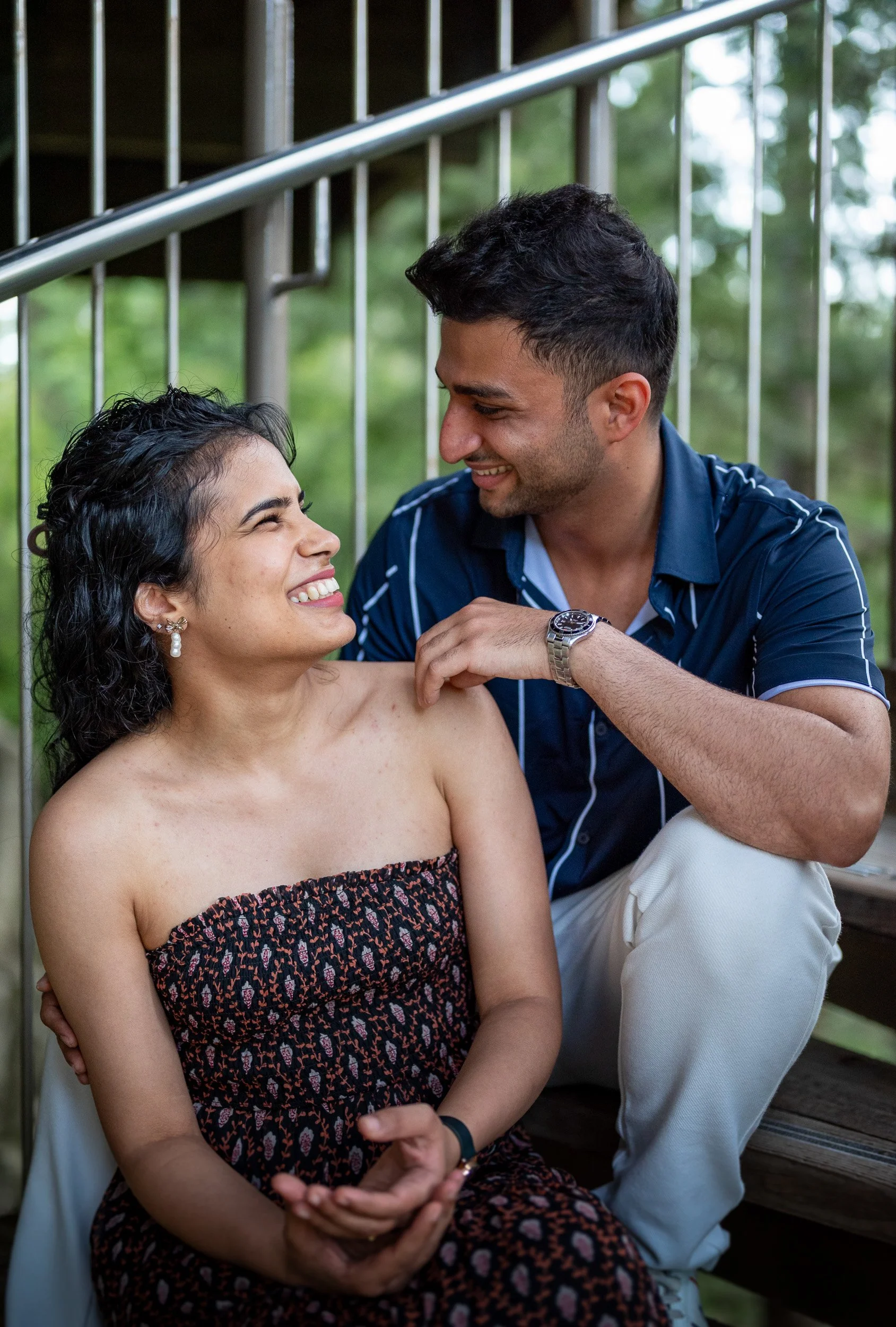 A smiling woman with dark, curly hair, wearing an off-shoulder dress, sitting next to a smiling man with short, dark hair, wearing a navy blue shirt, outdoors near a metal railing with green trees in the background.