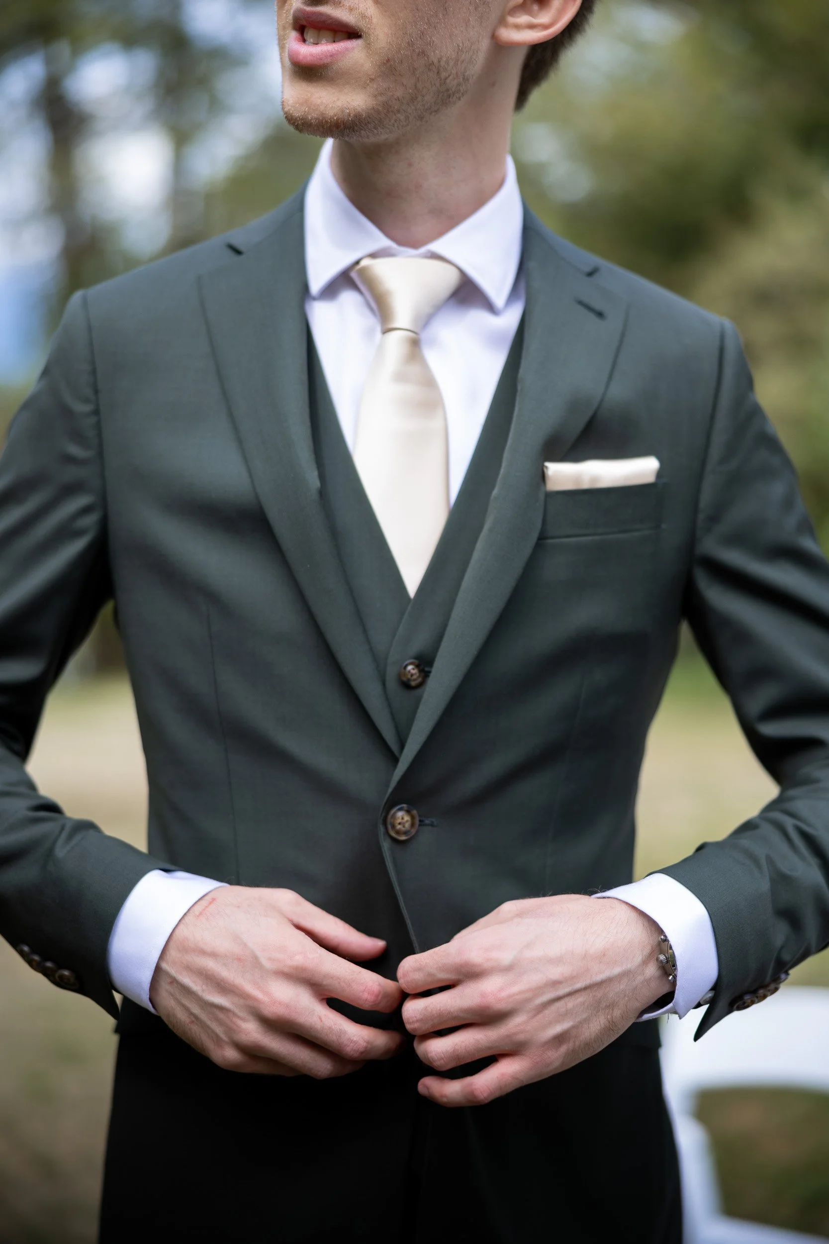 Close-up of a groom in a dark gray suit, white shirt, and cream-colored tie, standing outdoors with blurred trees in the background at Queen Elizabeth Park.
