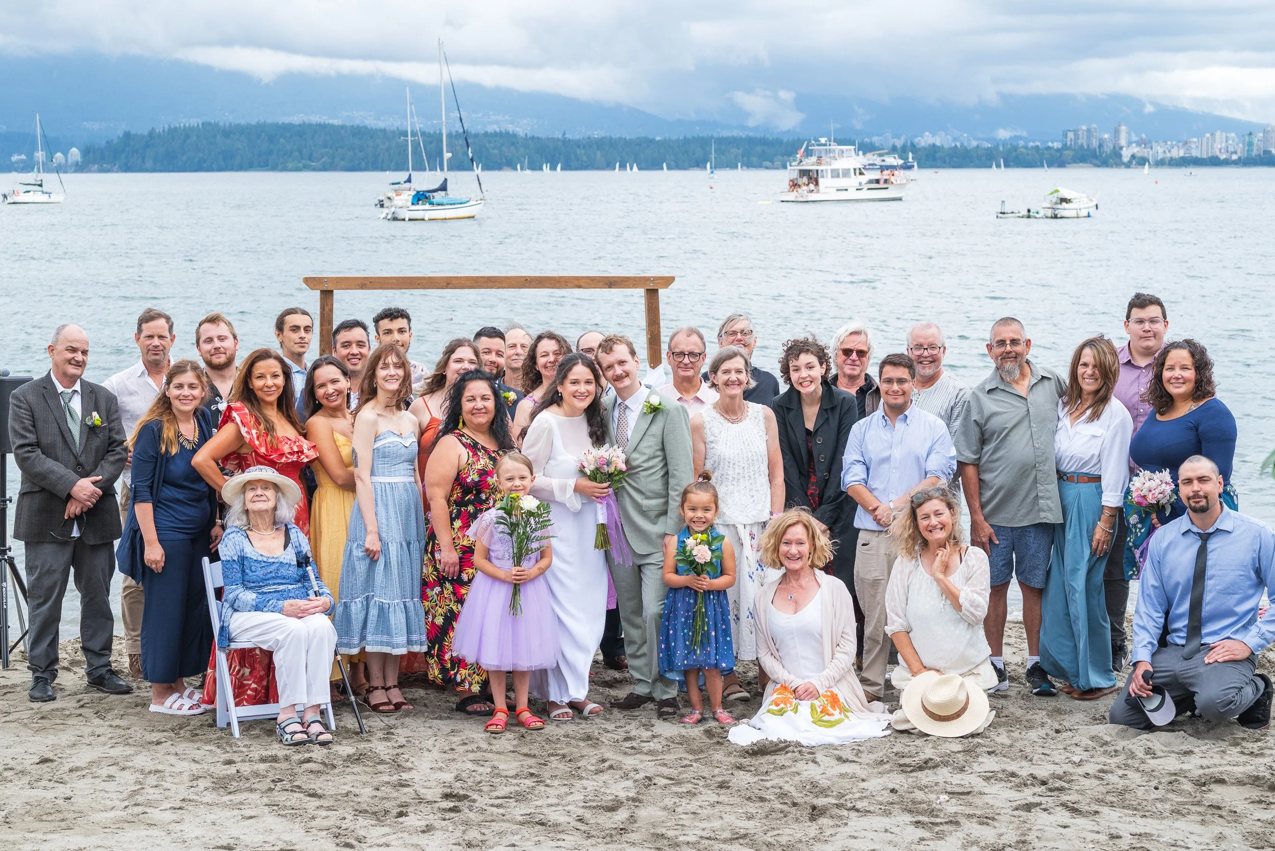 A large group of people, including children and adults, gathered on a sandy beach for a celebration or wedding, with sailboats and a city skyline in the background.