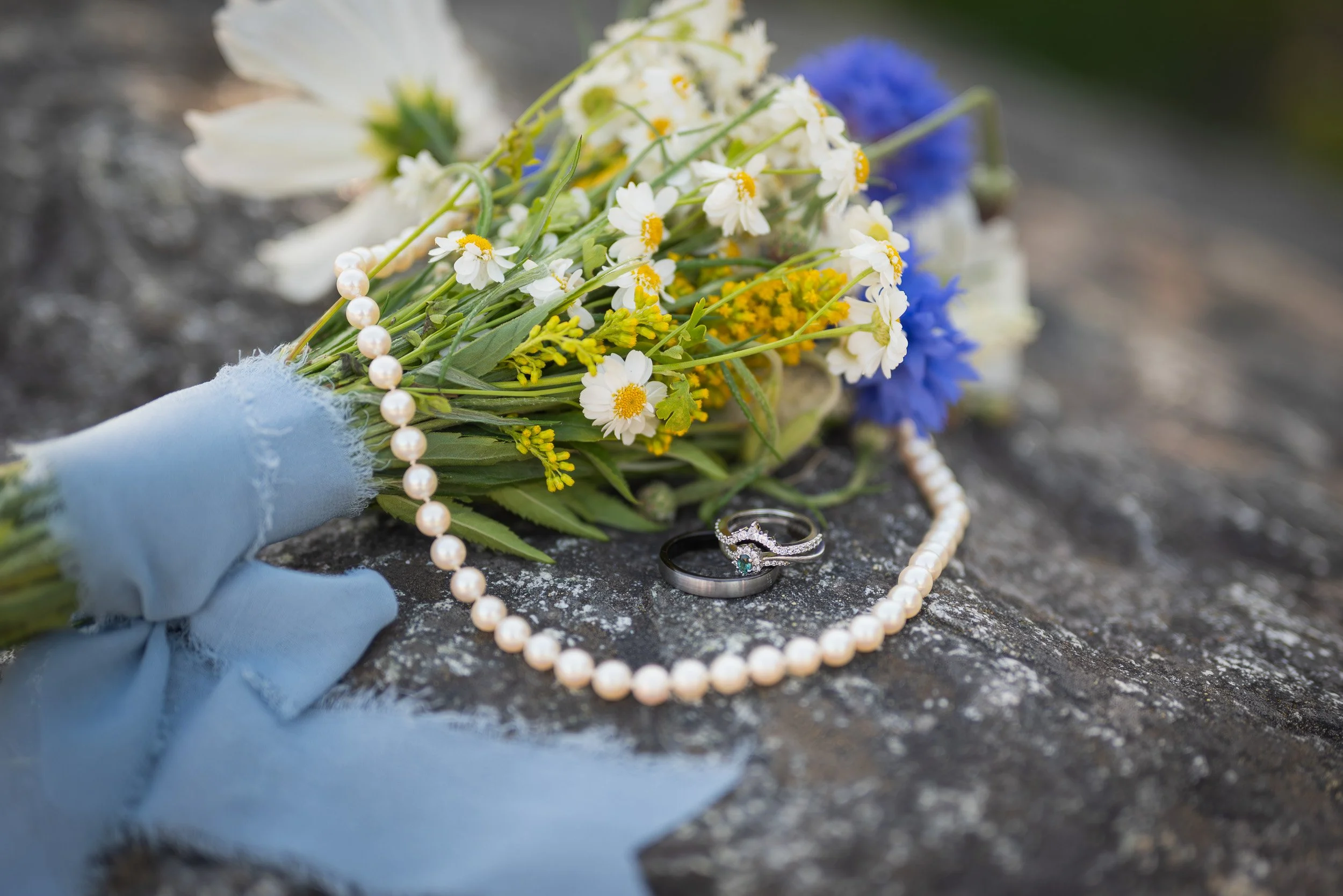 A small bouquet of white, yellow, and purple flowers lying on a dark stone surface, accompanied by a string of white pearls, a diamond engagement ring, and a wedding band, suggesting a memorial or tribute scene.