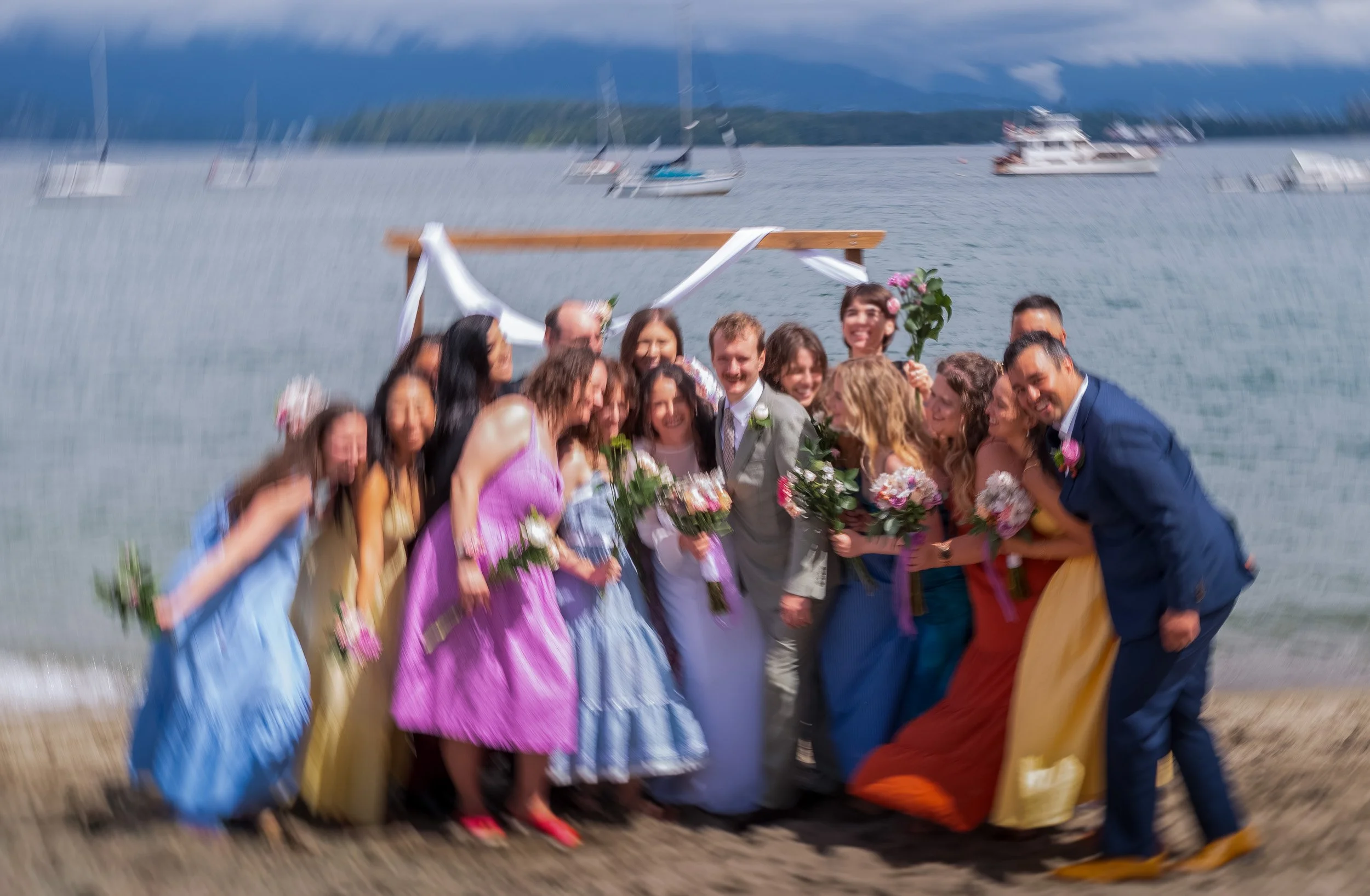 A group of people celebrating a wedding by a lake, with boats and sailboats in the background.