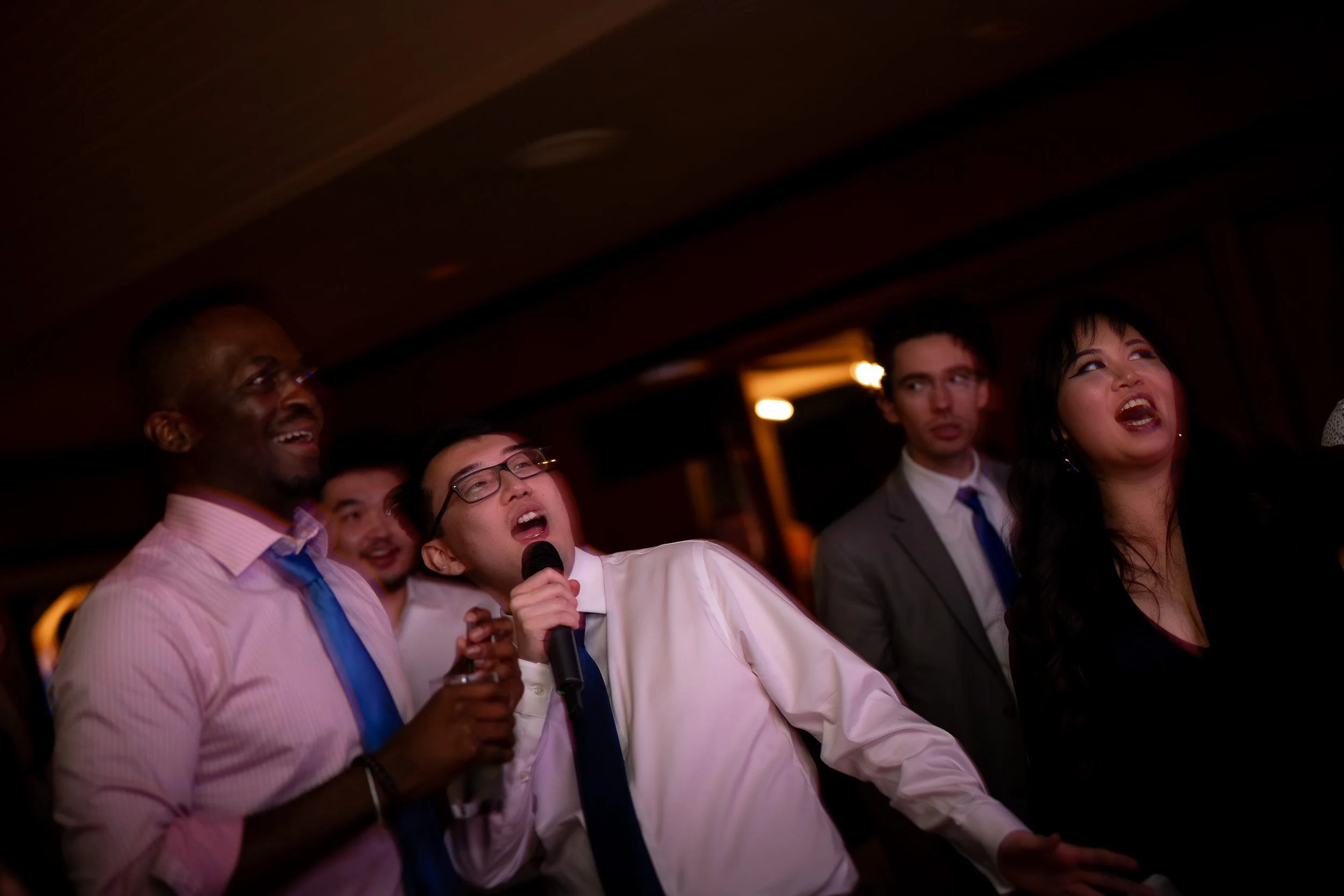 A diverse group of people at a social gathering, some smiling and one holding a microphone, in a dimly lit indoor setting.