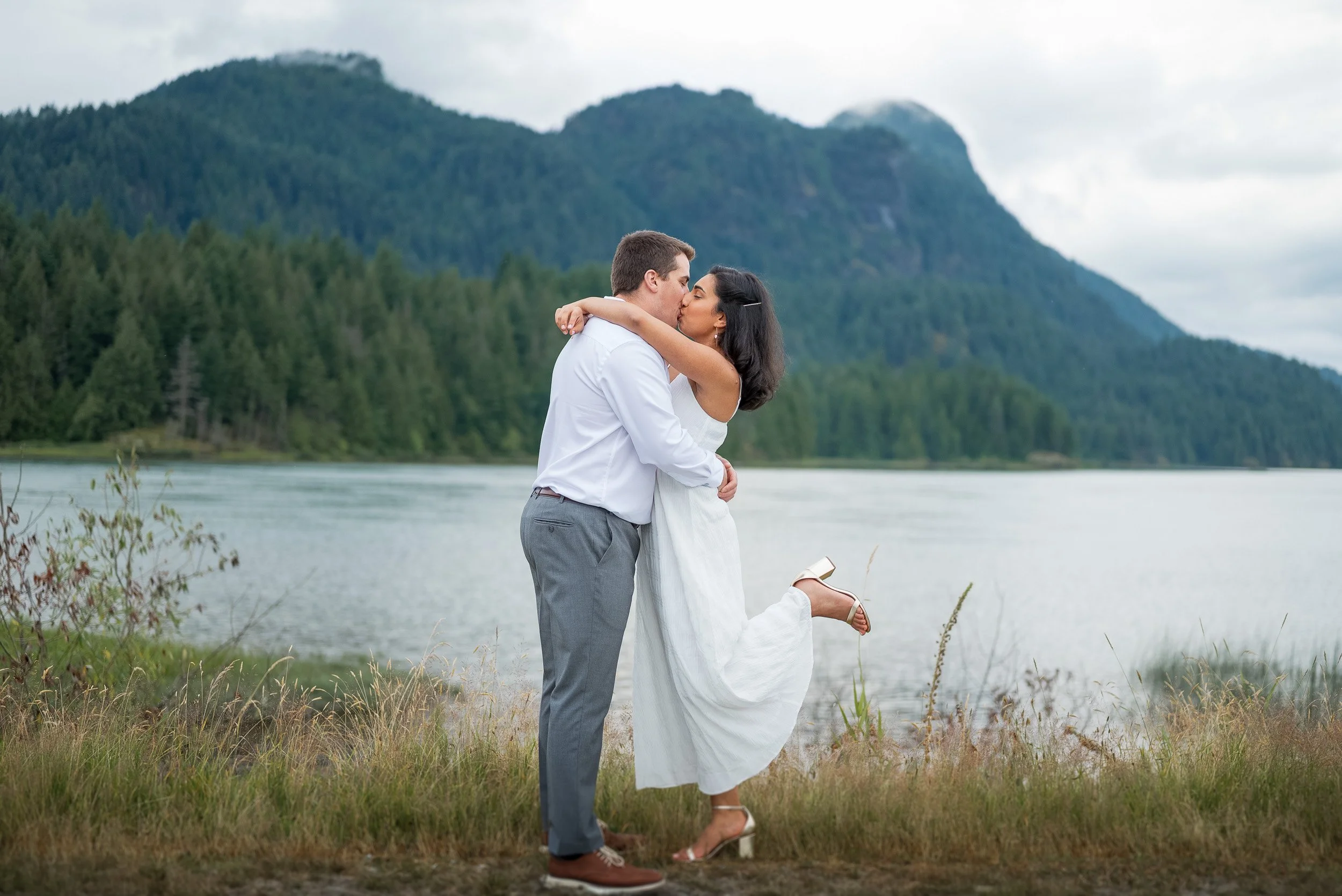 A couple embracing and kissing outdoors by a lake with a mountainous forested landscape in the background at Pitt Lake in Pitt Meadows.