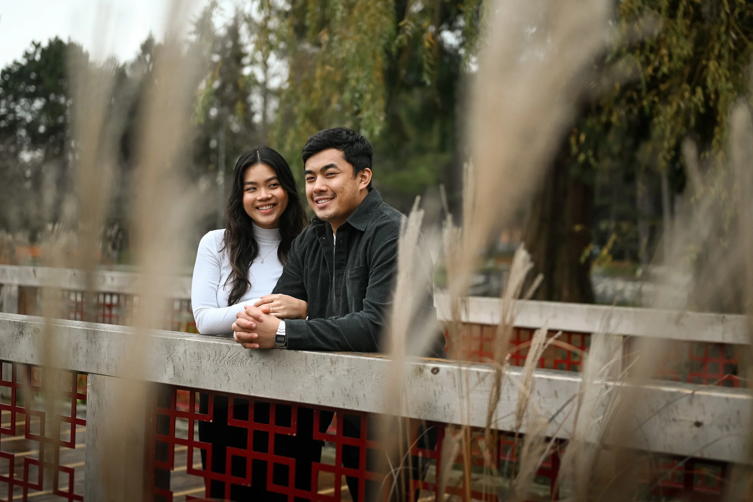 A photoshoot of a smiling young couple sitting outdoors behind a wooden fence with greenery in the background, viewed through dried grasses at Minoru Park in Richmond, BC