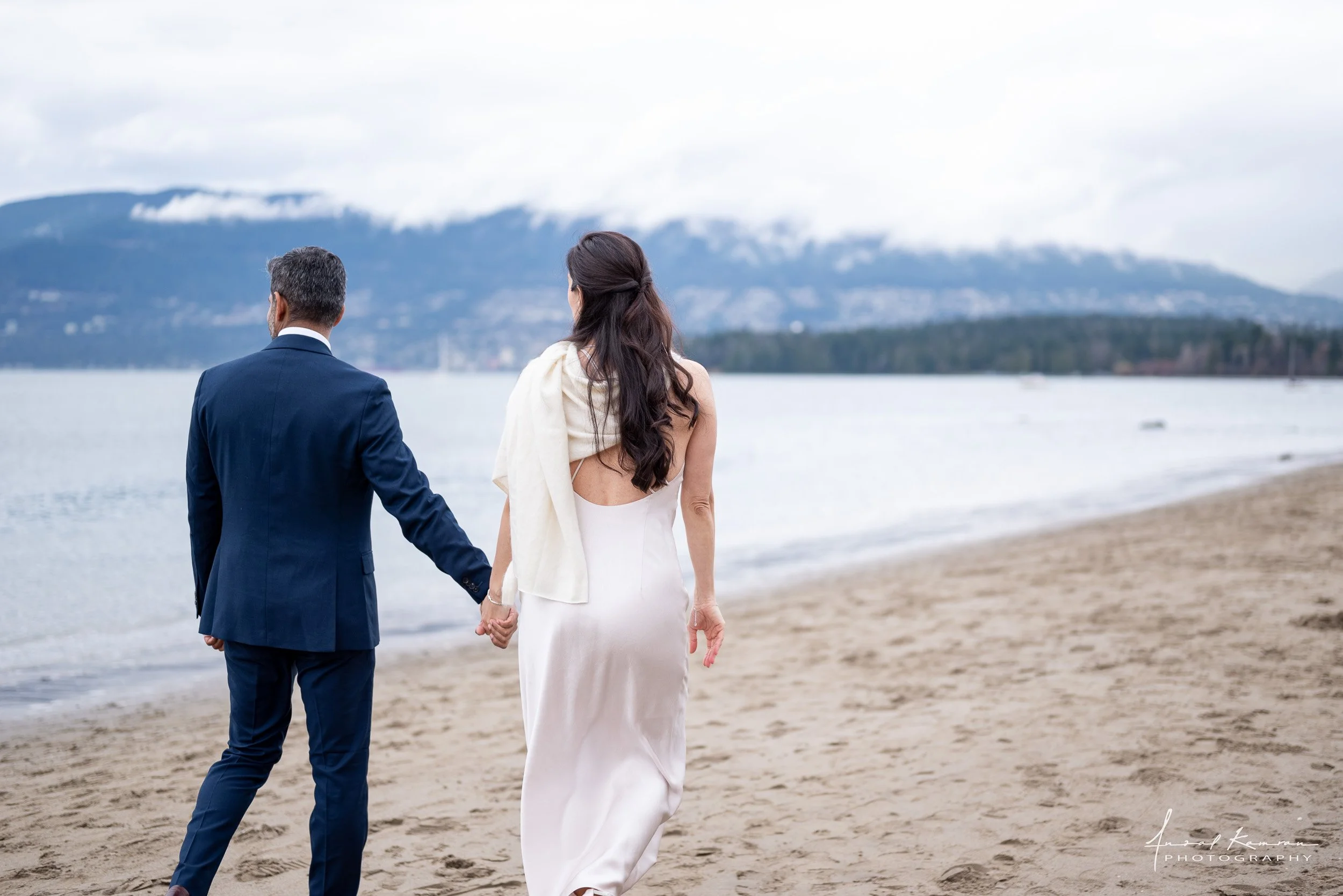 A couple dressed in wedding attire holding hands and walking along a beach, with mountains and cloudy sky in the background.