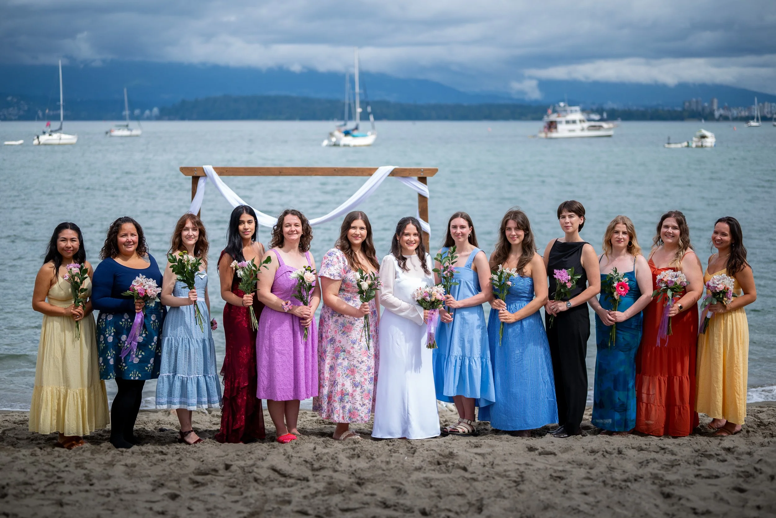 A group of bridesmaids and bride dressed in colorful dresses, holding flowers, standing on a beach at Jericho Beach Vancouver Wedding with sailboats and a boat in the water in the background.
