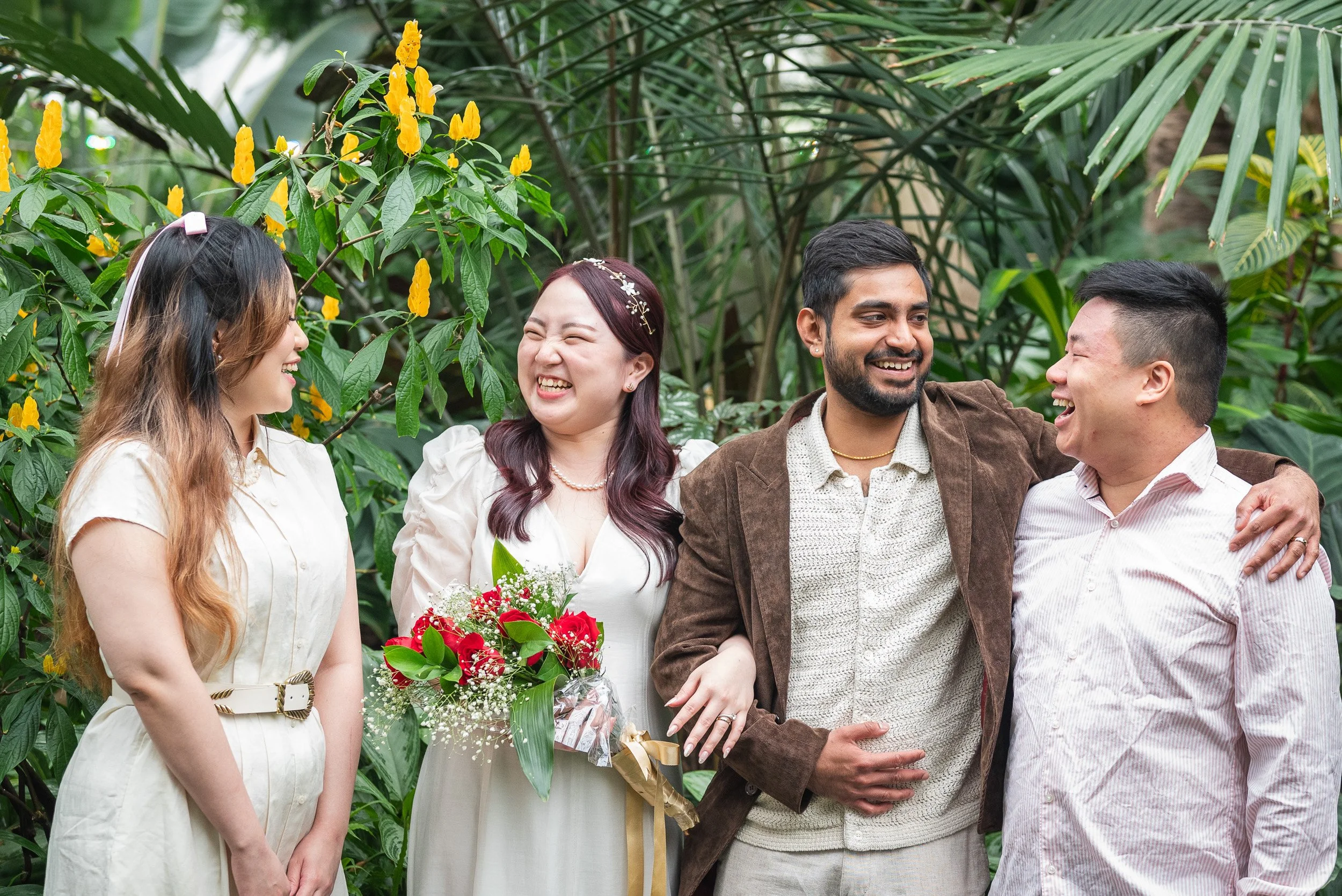 A group of five people smiling and chatting in a lush garden, with a woman holding a bouquet of red and white flowers.