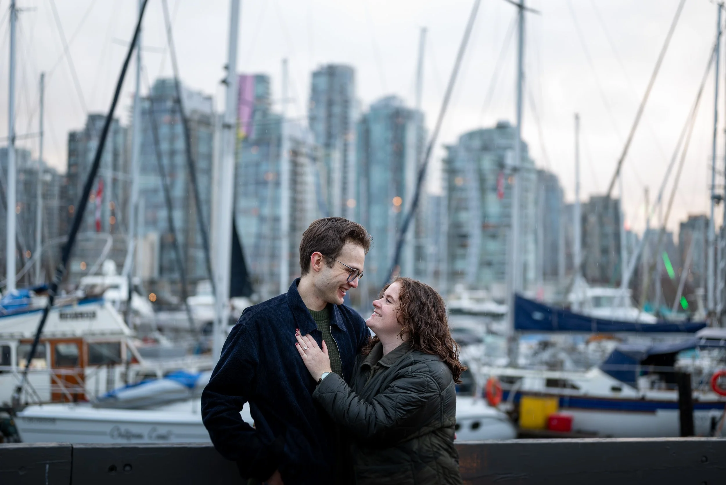 A smiling couple standing close together at a marina with boats and Vancouver city high-rise buildings in the background.