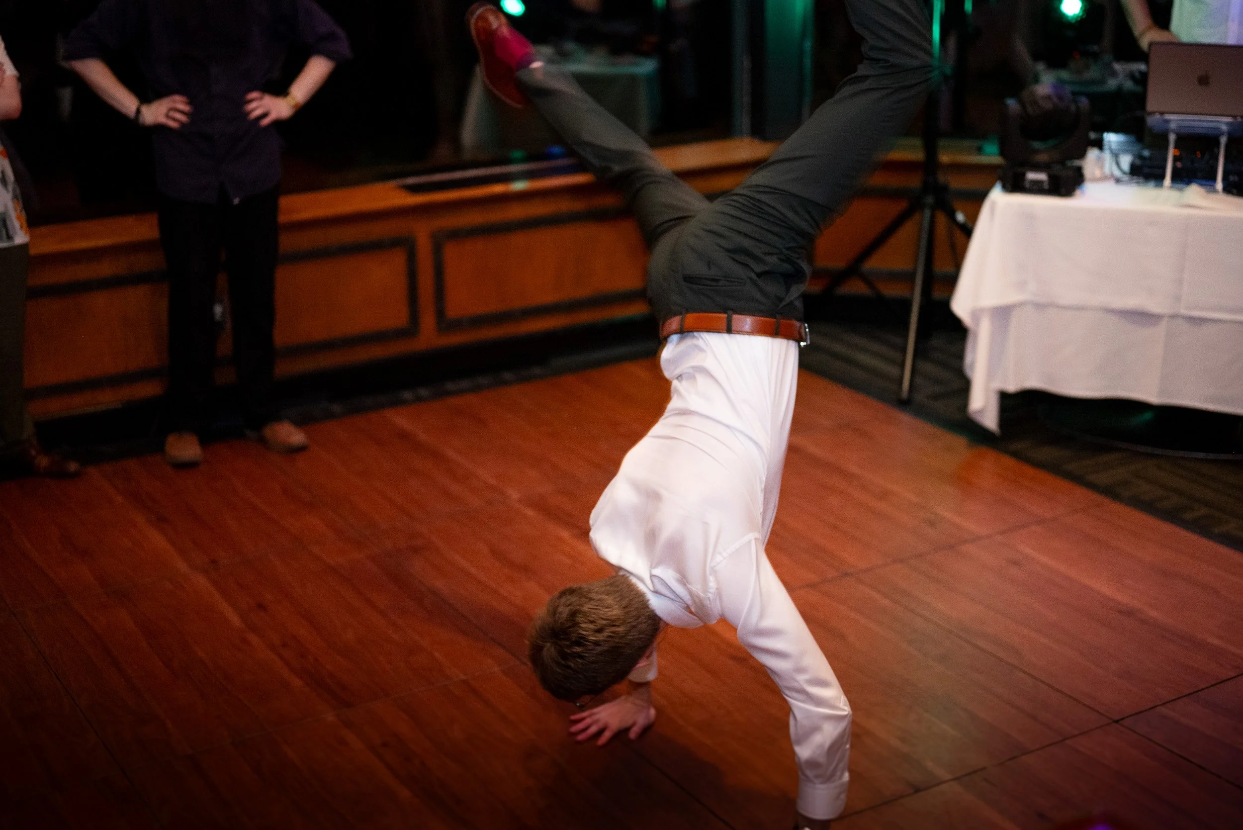 Man performing a handstand on a wooden dance floor with hands on the ground and feet in the air, while others watch, in a dimly lit room.