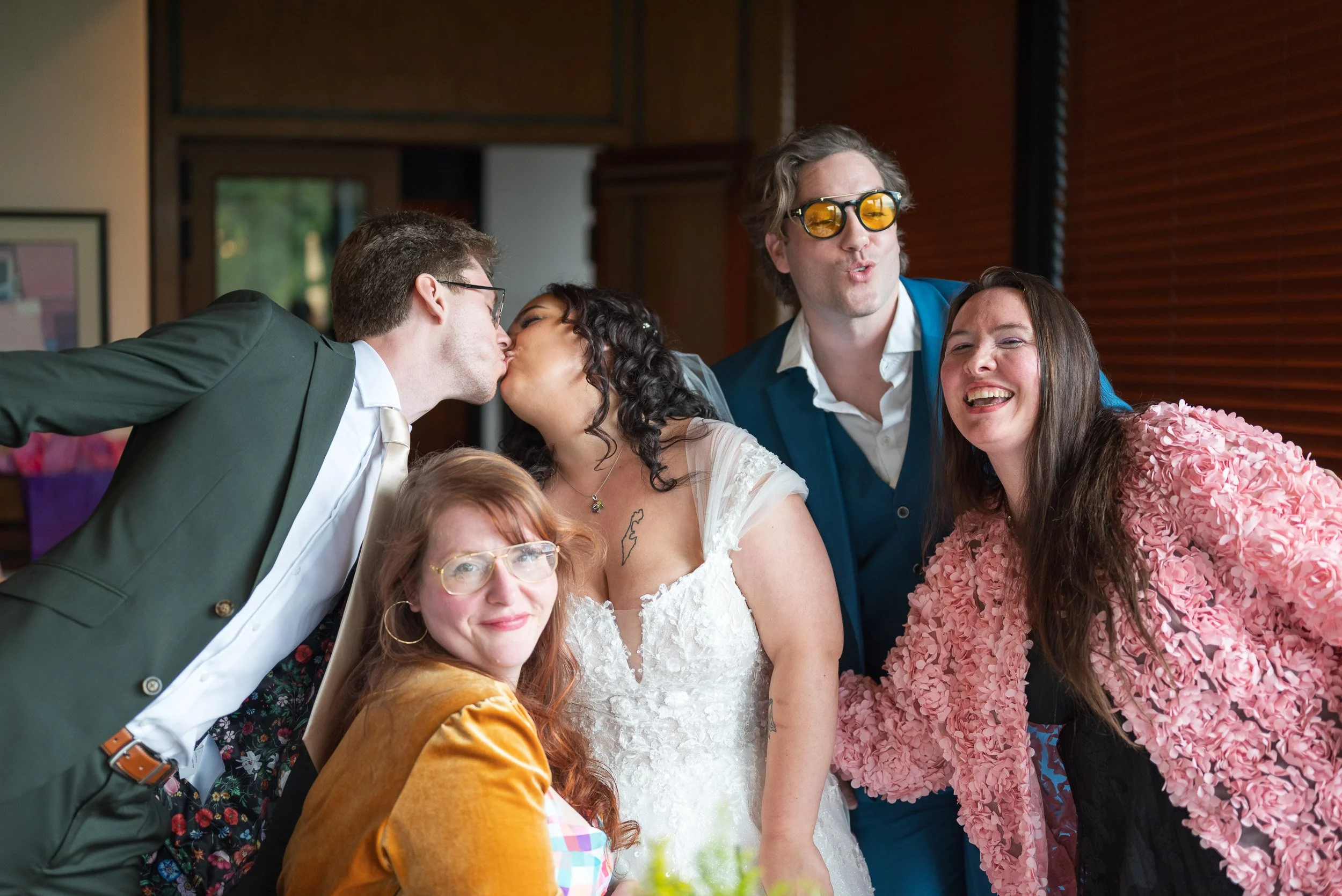 Group of people at a wedding, with the bride and groom kissing, and others smiling and posing for photos.