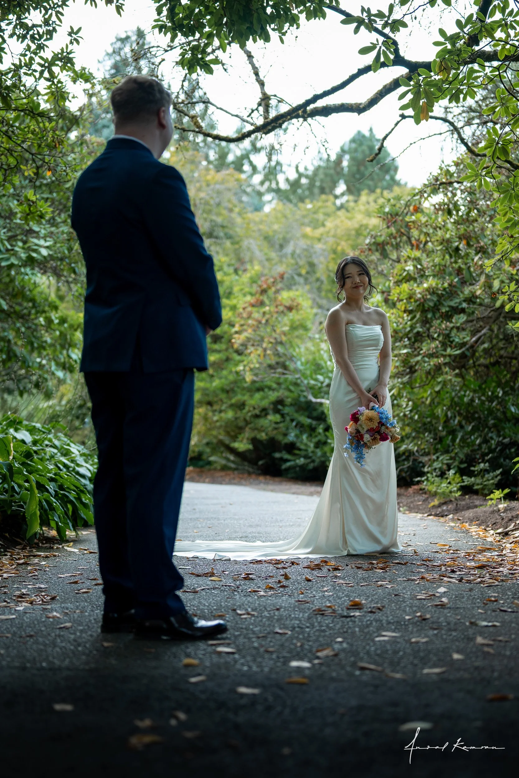 A bride holding a colorful bouquet of flowers is looking at a groom dressed in a dark suit outside surrounded by greenery.