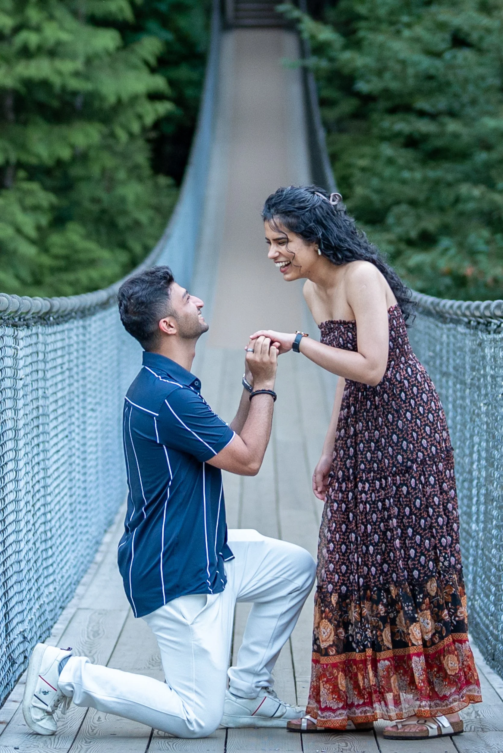 A man kneeling on one knee proposing to a woman on Capilano Suspension bridge surrounded by green trees. The woman is smiling and holding the man's hands.