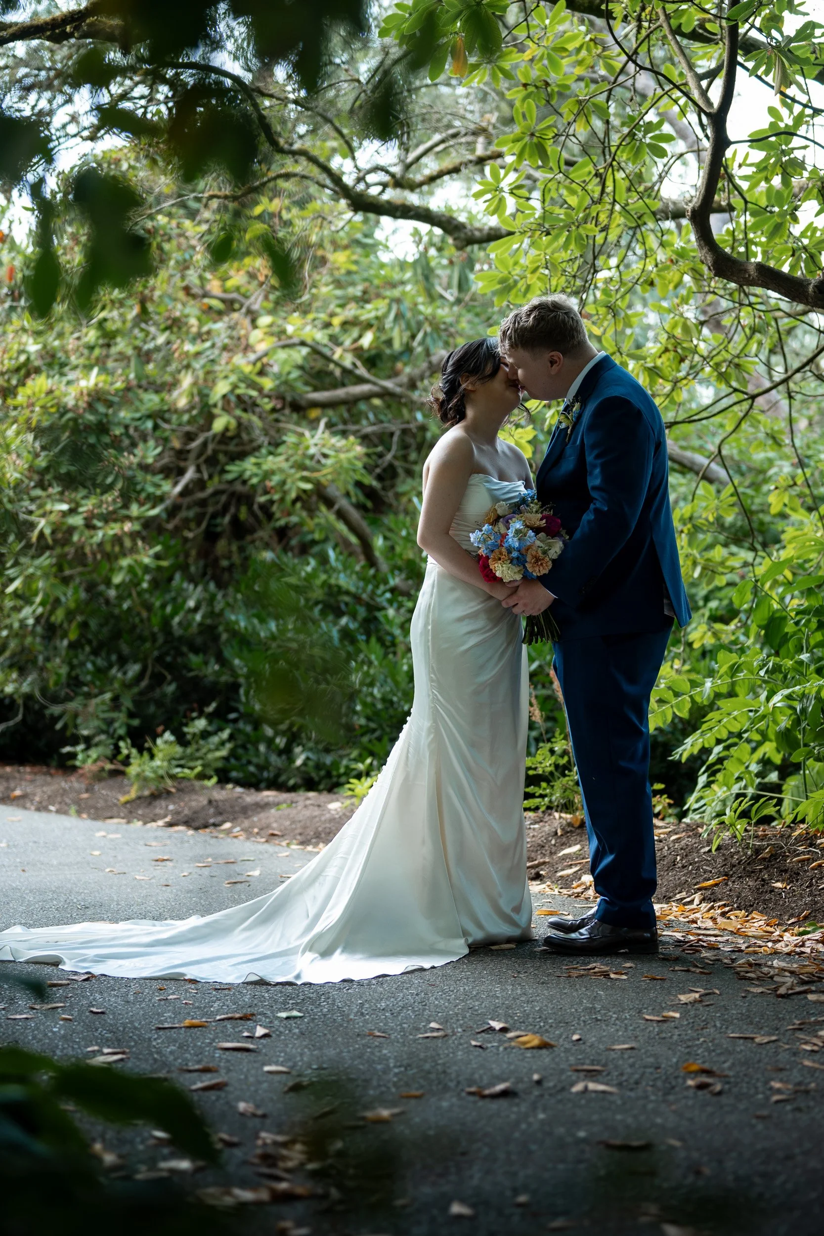A bride and groom are standing close together in a lush, green outdoor setting, sharing an intimate moment with their foreheads touching. The bride is holding a colorful bouquet, wearing a strapless white wedding gown, and the groom is dressed in a dark blue suit with a boutonniere. They are surrounded by trees and foliage, creating a natural and romantic atmosphere.
