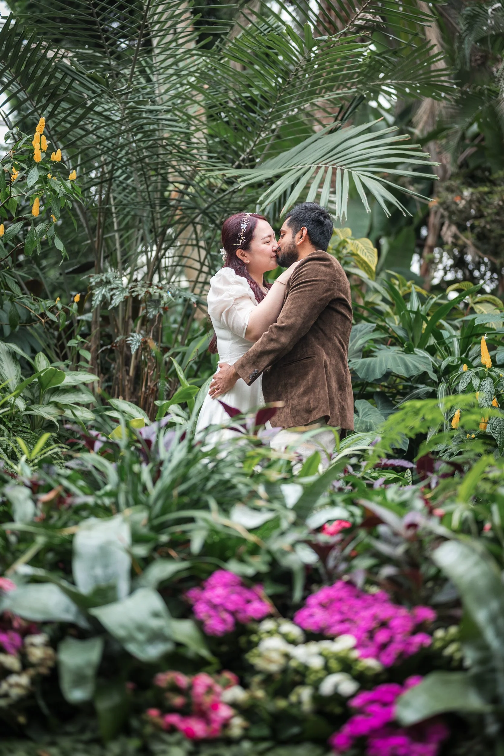 A couple kissing in a lush, tropical garden surrounded by large green leaves and colorful flowers at Bloedel Conservatory