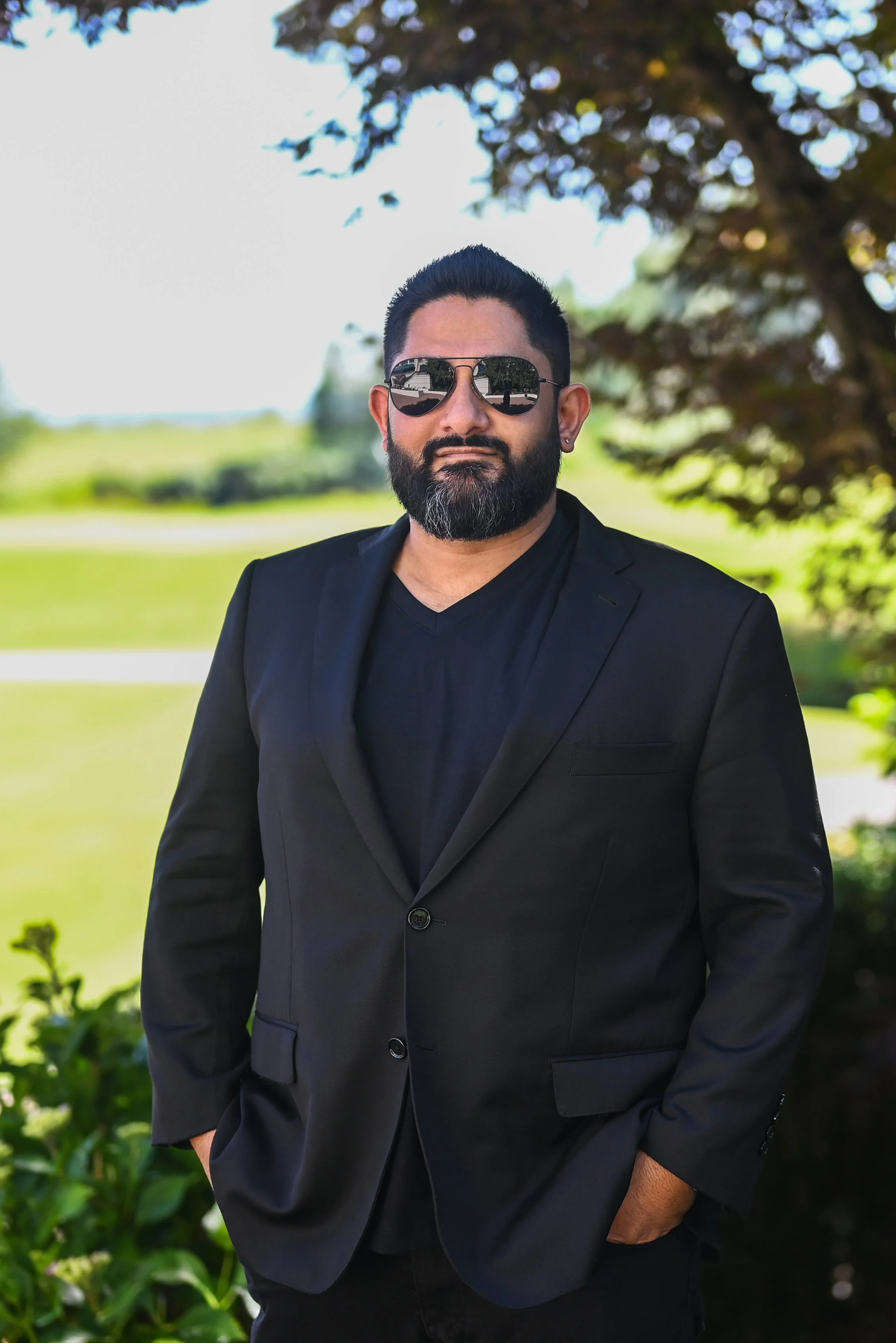 A man with dark hair and a beard wearing sunglasses, a black blazer, and a black shirt stands outdoors near green bushes and trees, with a blurred background of a park or garden. This is Anzal Kamran, a wedding and proposal photographer in Vancouver.