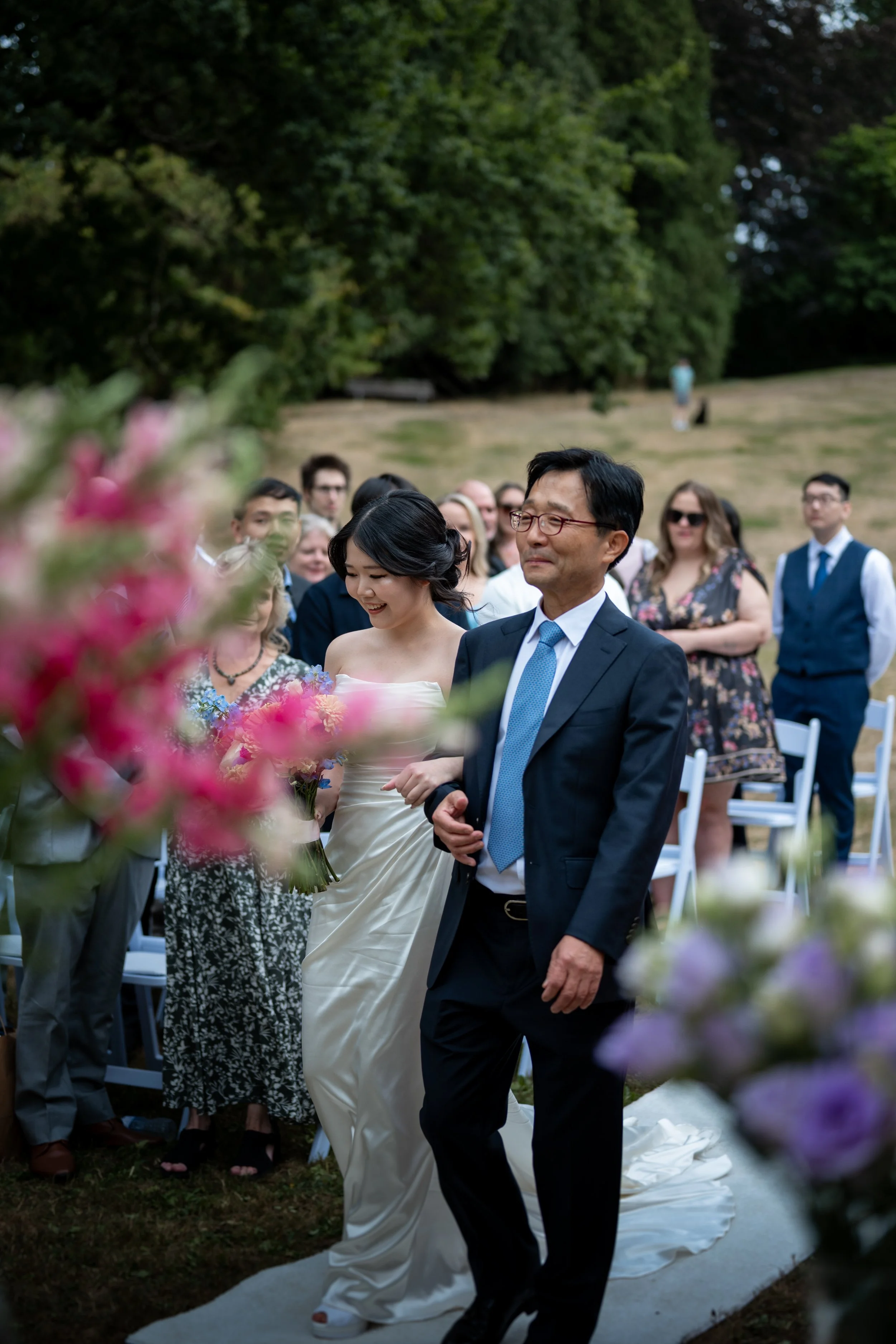 A bride and her father walking down the aisle at an outdoor wedding ceremony, surrounded by guests and greenery at Queen Elizabeth Park Duck Pond.