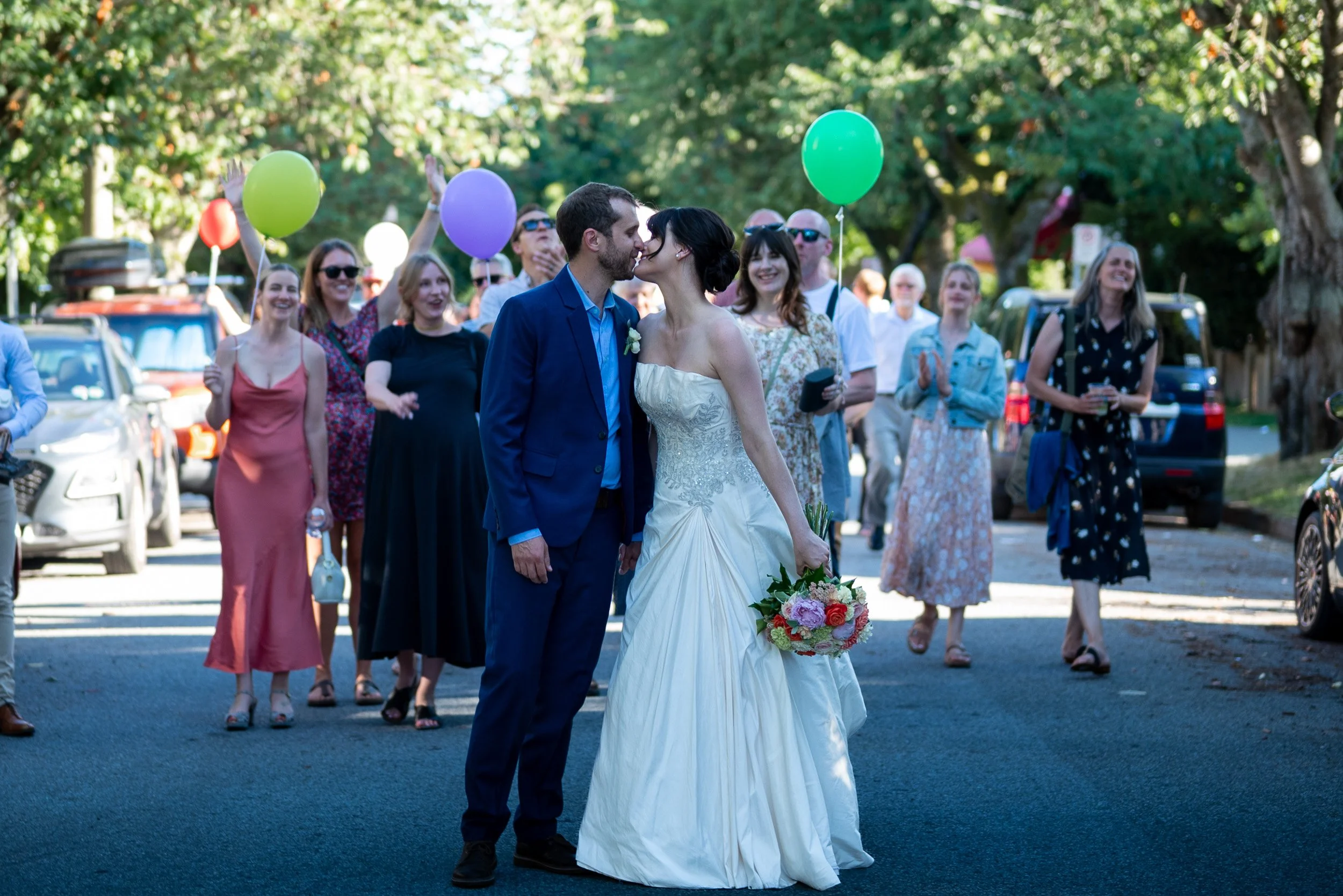 A bride and groom kissing at their wedding surrounded by guests, some holding balloons, outdoors with trees and parked cars in the background on the street of Vancouver