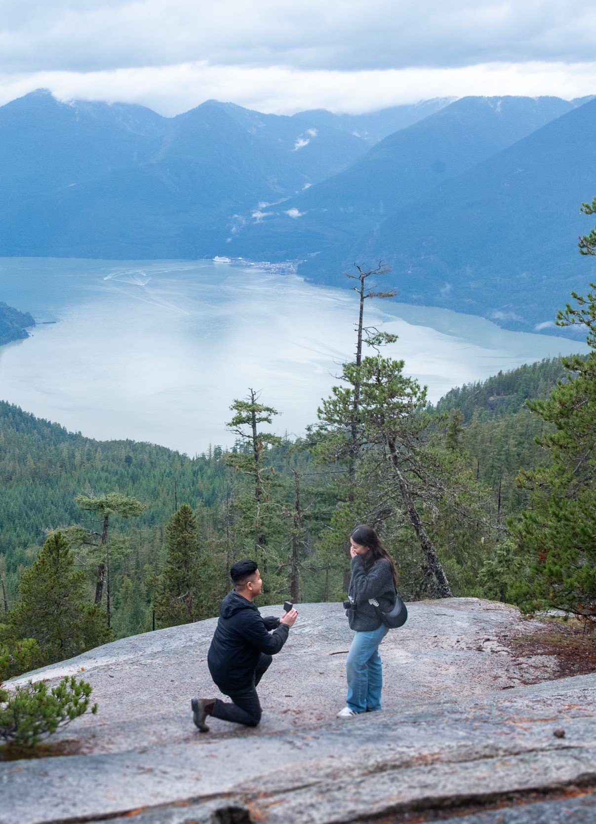 A man proposing marriage to a woman on a large flat rock in a scenic mountainous area with a lake and forest in the background at Sea to Sky Gondola.