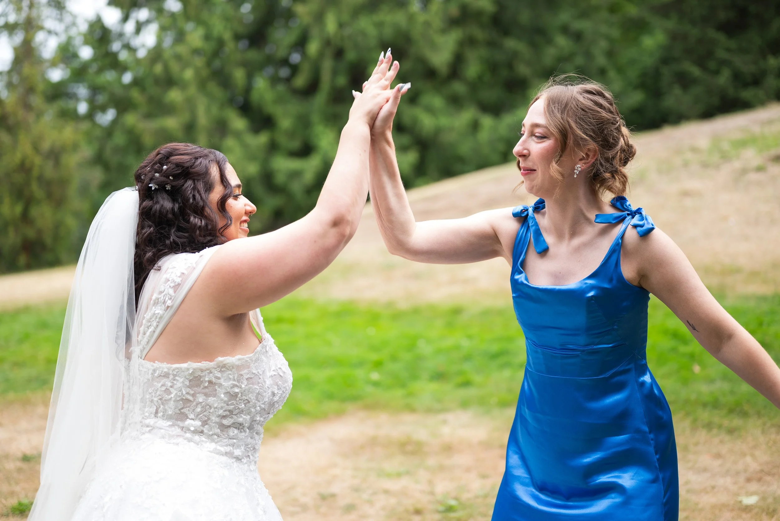 Two women, one in a wedding dress and veil, and the other in a blue dress, high-five outdoors with greenery in the background.